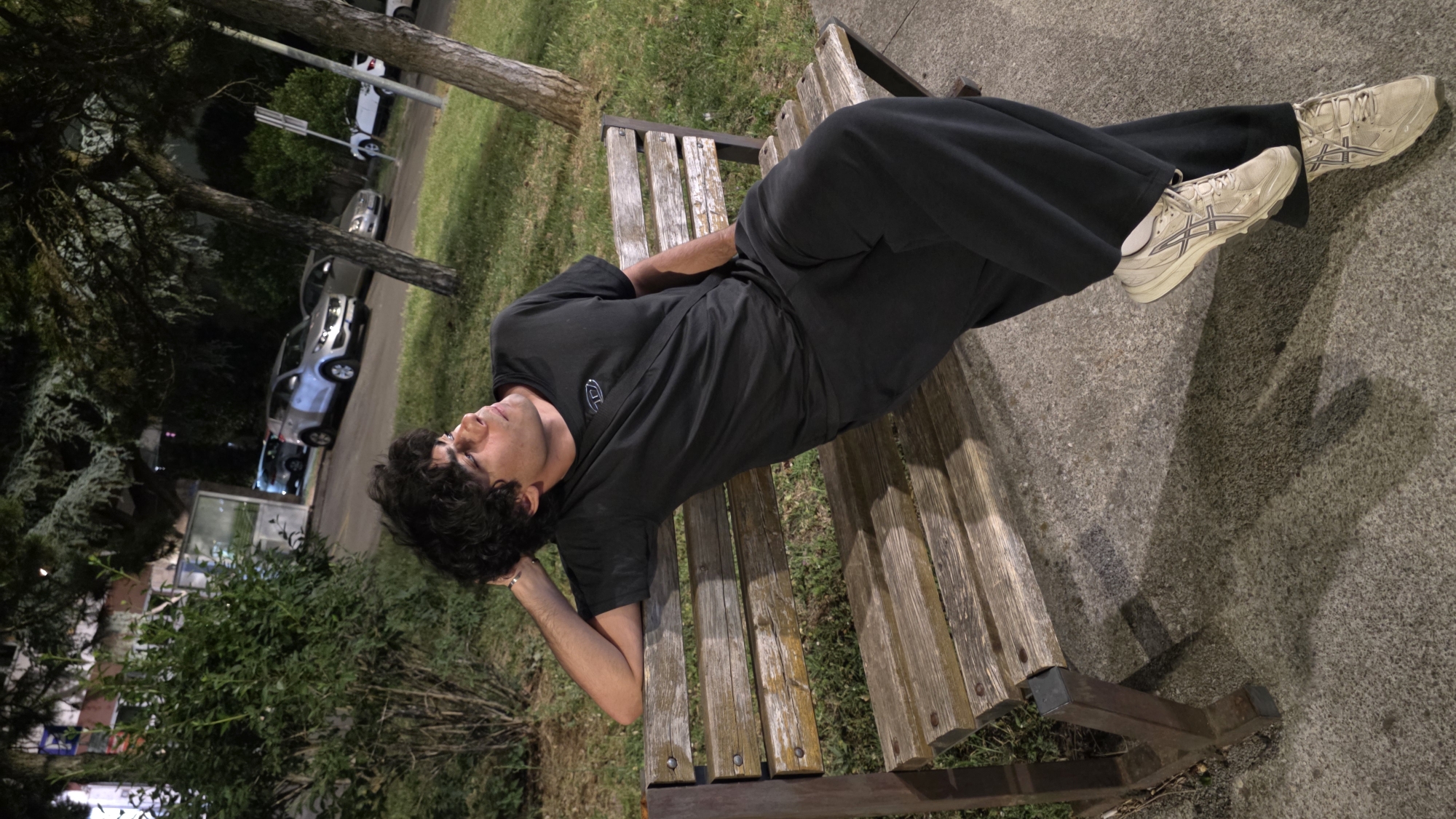 A young man is lying on a wooden bench outdoors. He is wearing a black t-shirt, black pants, and off-white athletic shoes. The bench is located on a paved surface next to a grassy area with trees and shrubs. In the background, there is a street with parked cars. The location is likely a park or public area in the city of Podgorica, Montenegro, based on the architecture and environment. The man appears to be resting or sleeping.