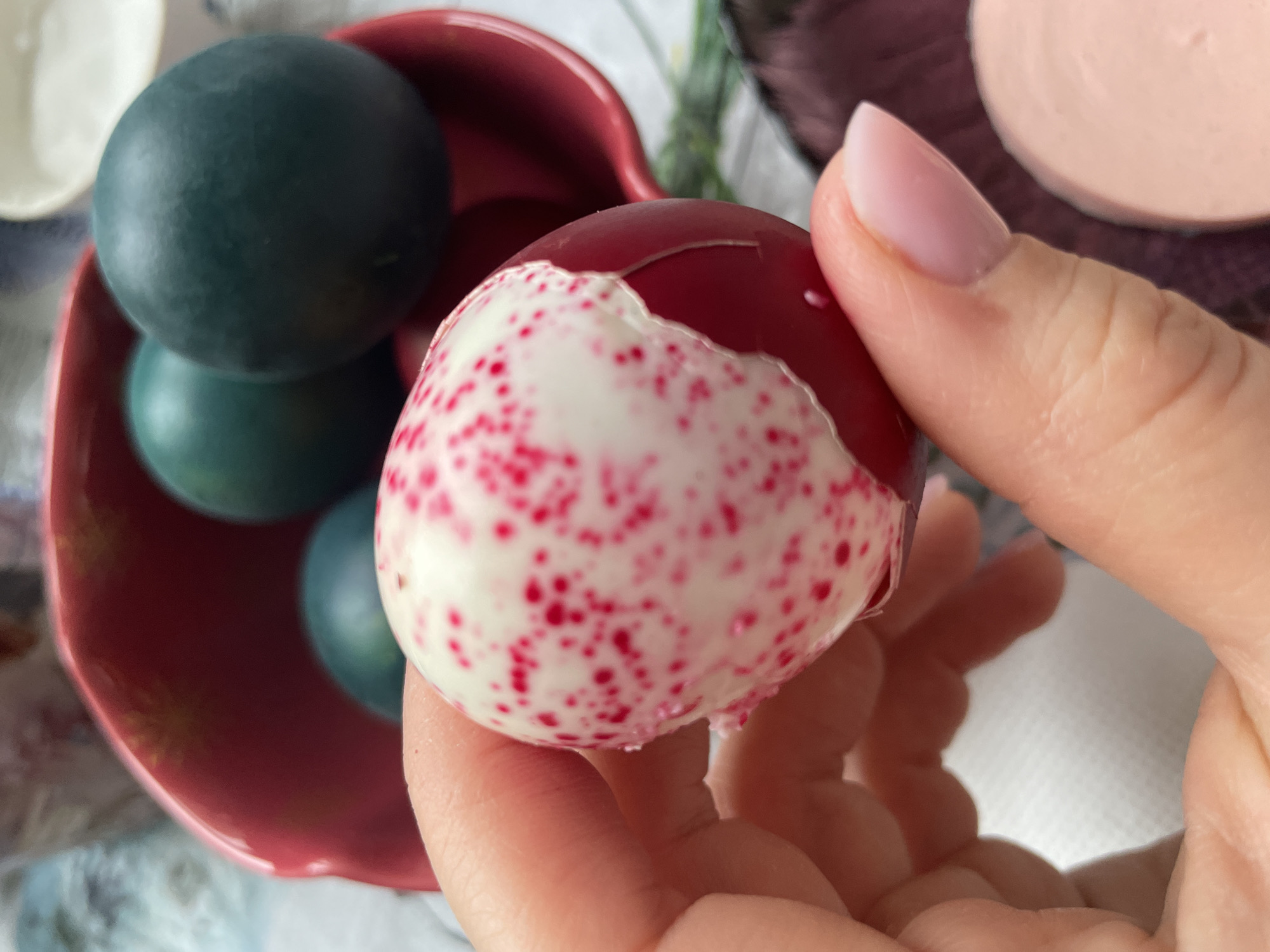 The image shows a person holding a dyed Easter egg with a cracked shell in their hand. The egg is mostly white with red speckles, and the exposed portion shows a solid red dye. The person's hand is visible, including a pink nail. In the background, there's a red bowl containing green dyed eggs. Another disc-shaped object is visible in the top right corner, possibly another egg or a decoration. The context and location mentioned suggest that the scene involves preparing for or celebrating Easter in Chaikovsky, Russia.