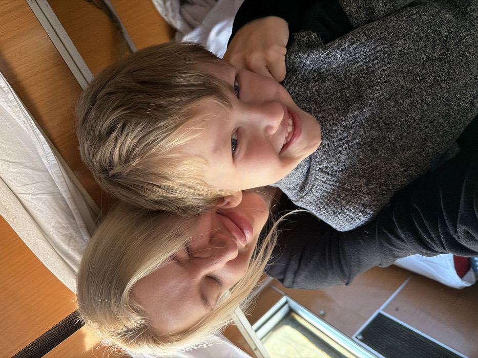 The image depicts an adult female and a young male child in a close-up, head-and-shoulders composition, set in an interior location identified as Bistrice, Montenegro. The female, with blonde hair, is positioned behind and to the left of the child. Her eyes are closed, and a subtle smile is present on her lips. She wears a dark, long-sleeved garment. Her right arm is visible, extended around the child's shoulder area. The child, with light brown hair, is in the foreground, facing the camera with a wide smile, displaying visible teeth. He wears a grey, ribbed knitted sweater. The background consists of light brown wooden paneling. In the upper right, white, crumpled fabric, possibly a curtain or bedding, is visible. A window frame is partially visible in the lower-left background, revealing a blurred exterior with hints of light and natural elements. Below the window, a dark, rectangular metallic vent or grille is present. The interior design suggests a confined space, consistent with a vehicle cabin such as a train or recreational vehicle.