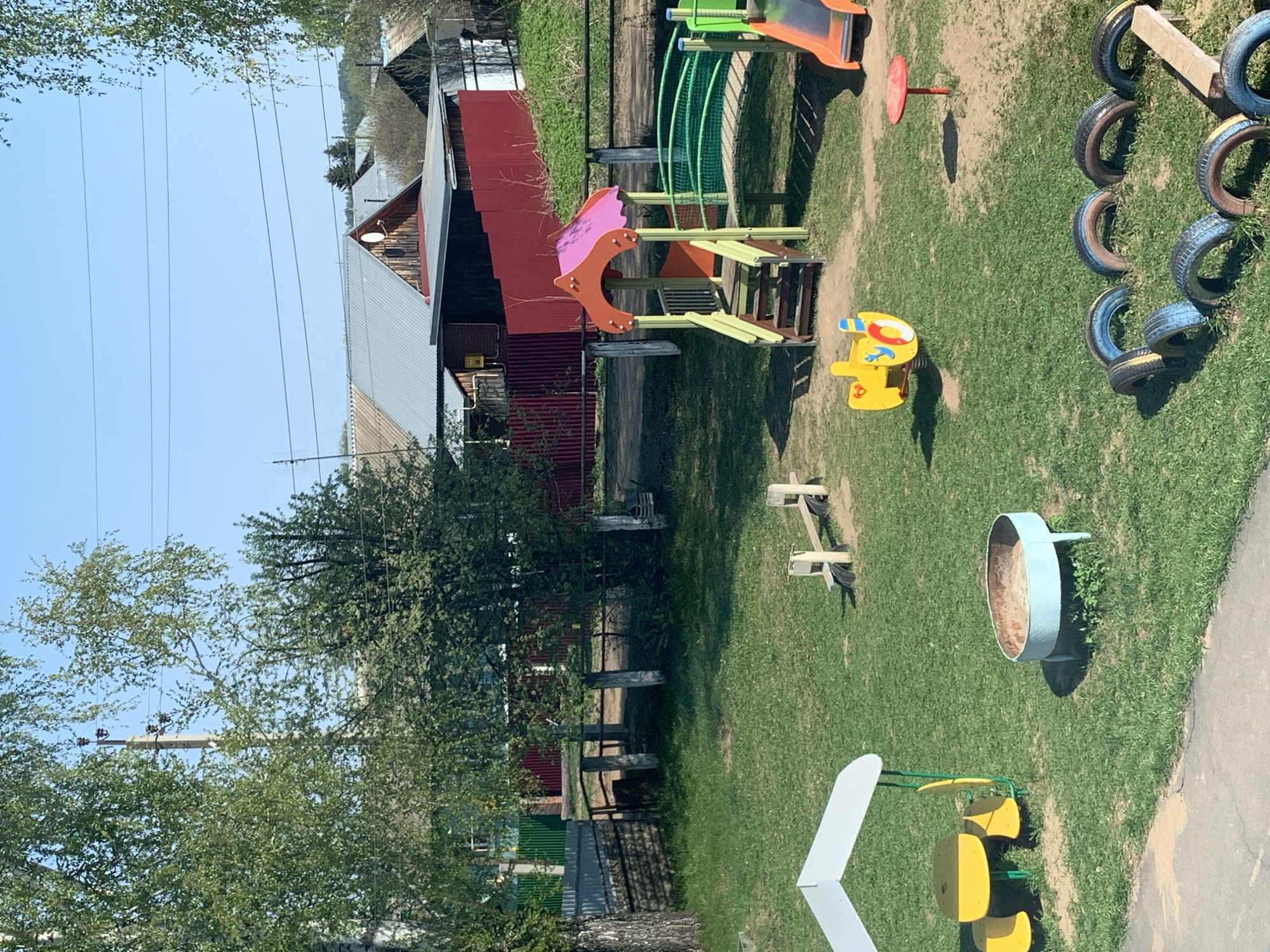 The image depicts a playground in Фоки, Russia. The playground features various equipment, including a slide, swings, a rocking horse, and structures made from tires. A building with a red exterior is visible in the background, adjacent to the playground. Utility poles with power lines are present, suggesting the playground is located in a developed area. The environment is characterized by greenery, indicating an outdoor setting. There are no people visible in the photograph.