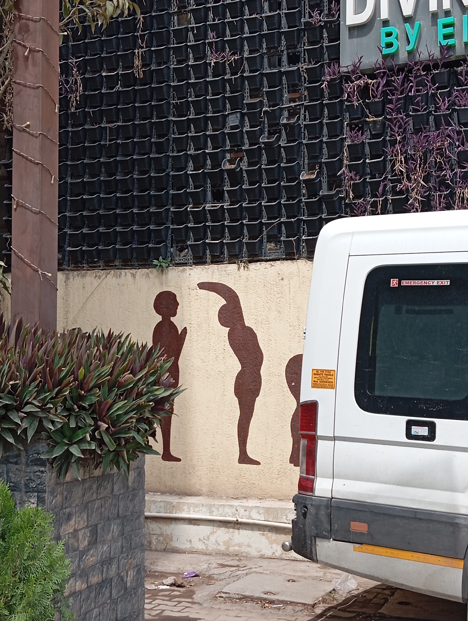 A partially obscured, medium-shot photograph depicts a section of a building wall in Jonk, India, adorned with stylized silhouettes of human figures in various poses, adjacent to a white van parked on a street.
The building wall, painted in a light beige color, showcases three brown silhouettes of human figures. The figure on the left appears to be in a standing position with hands held together in front of the chest. The central figure is depicted in a stretching pose, leaning backward with one arm raised overhead. The figure on the right is smaller and obscured. Above the wall, rows of stacked black planters create a vertical garden effect.
To the right, the rear portion of a white van is visible. Details like the brake light, bumper, and an "Emergency Exit" sticker on the rear window are discernible. A sticker with text in a local language is also noticeable.
In the foreground, a planter box constructed of dark gray stone holds a bush of green and purple plants. The street in front of the wall and van appears to be paved and has some debris. The overall scene suggests an urban location, potentially near a wellness center or yoga studio, as indicated by the figures on the wall. The image is taken during daylight hours.