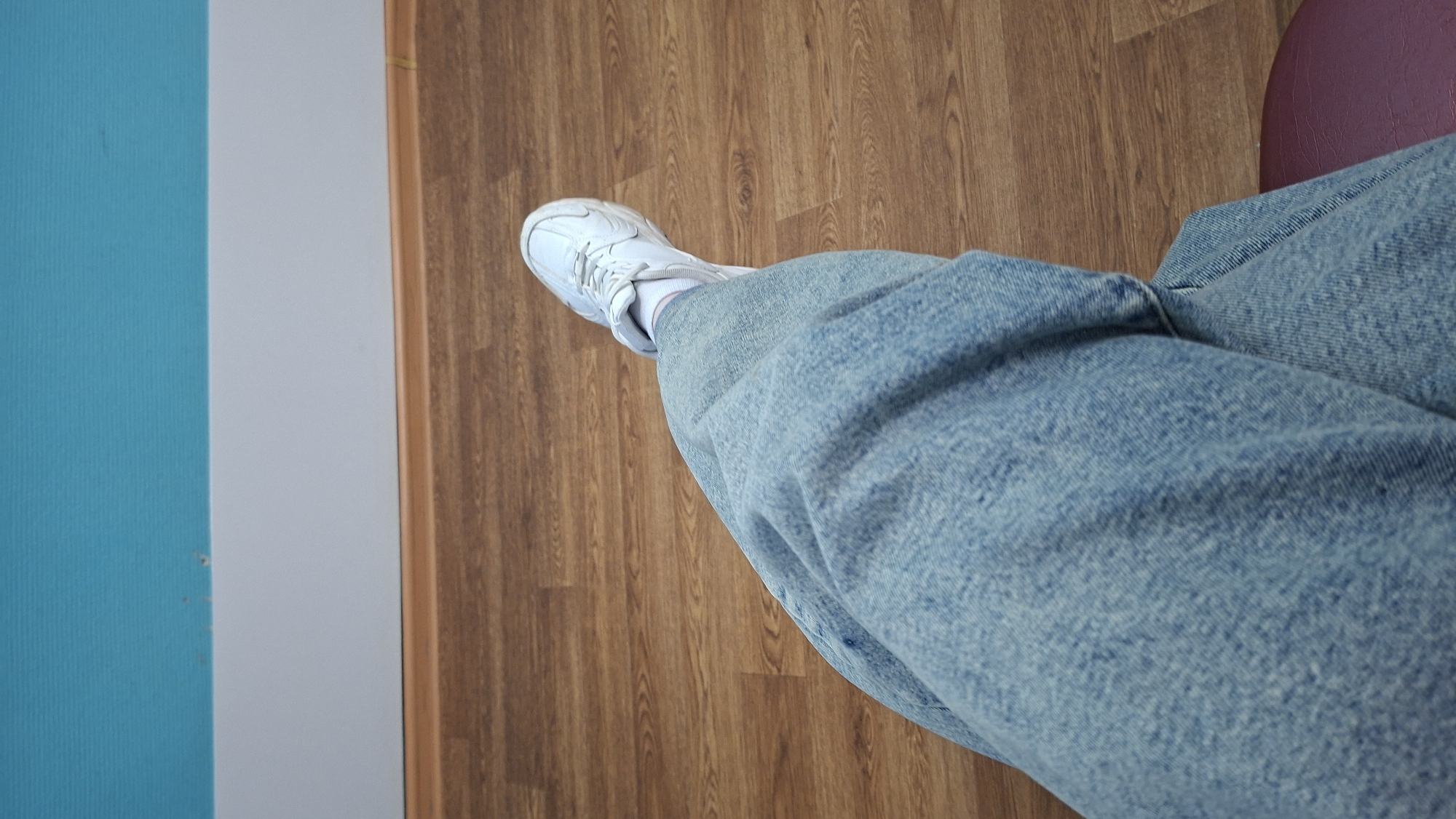 The image displays a high-angle shot looking down at a person's lower leg and foot within an indoor setting in Chaikovsky, Russia. The subject is wearing light blue, possibly wide-leg, denim jeans and a white sneaker with white laces. White socks are visible between the shoe and the hem of the jeans.
The person appears to be standing or sitting, with their leg bent and positioned on a warm-toned wooden floor that covers most of the background. To the left, there is a wall divided into two sections: a textured turquoise wall on the far left and an adjacent light gray wall. A thin brown wooden trim or frame separates these wall sections from the wooden floor. The light gray wall shows a few minor scuff marks. In the upper right corner, a small, blurred segment of a purple object, likely part of a chair or cushion, is visible. The scene suggests a casual indoor environment, possibly a home or public building. No specific activity or emotions are discernible, and there is no visible text.