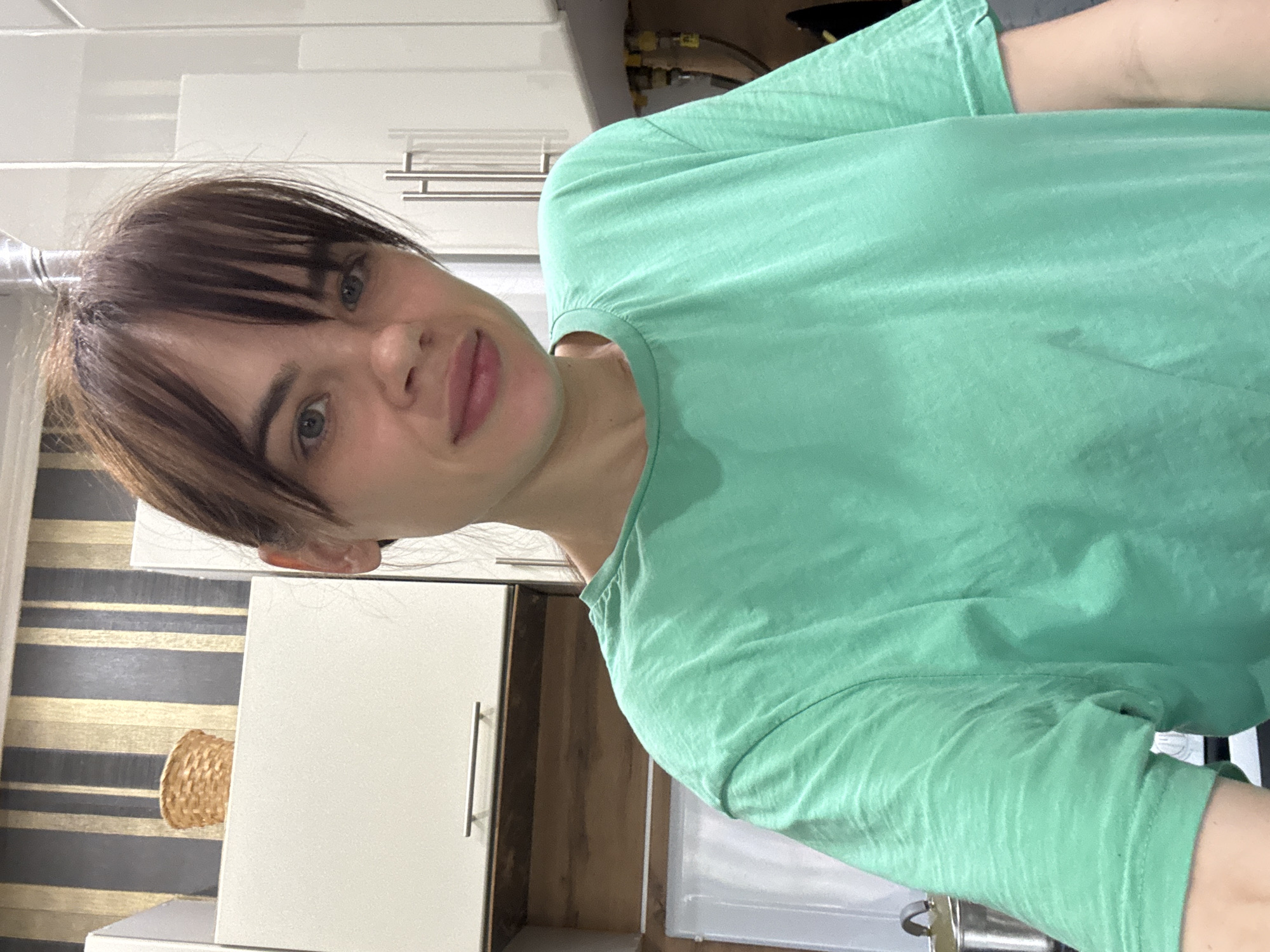 A person with dark hair and bangs is wearing a light green t-shirt and smiling at the camera. The person is indoors, seemingly in a kitchen, as evidenced by white cabinets and what appears to be a sink in the background. There is a woven basket visible on a shelf, and a patterned wall is behind the cabinets. The image does not provide any information about the location being Rostov-on-Don, Russia.