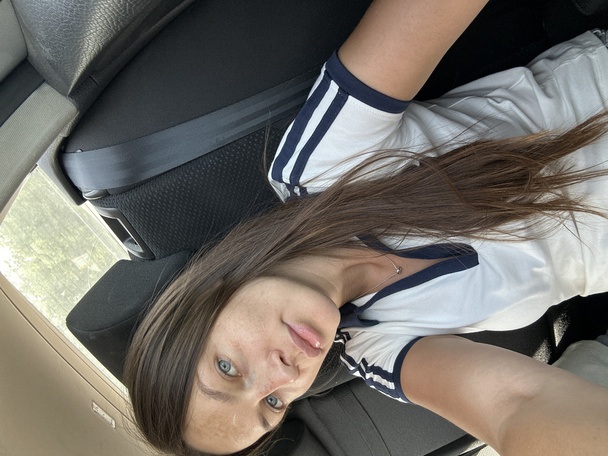 A woman with brown hair and blue eyes is visible in an interior car setting. She is wearing a white T-shirt with navy blue stripes on the sleeves and a delicate silver necklace. Her complexion exhibits some freckling. The interior of the vehicle is composed of dark grey seats and seat belts. Outside the car window, a glimpse of trees can be observed. The location in the city of Walsrode, Germany, cannot be determined from this image.