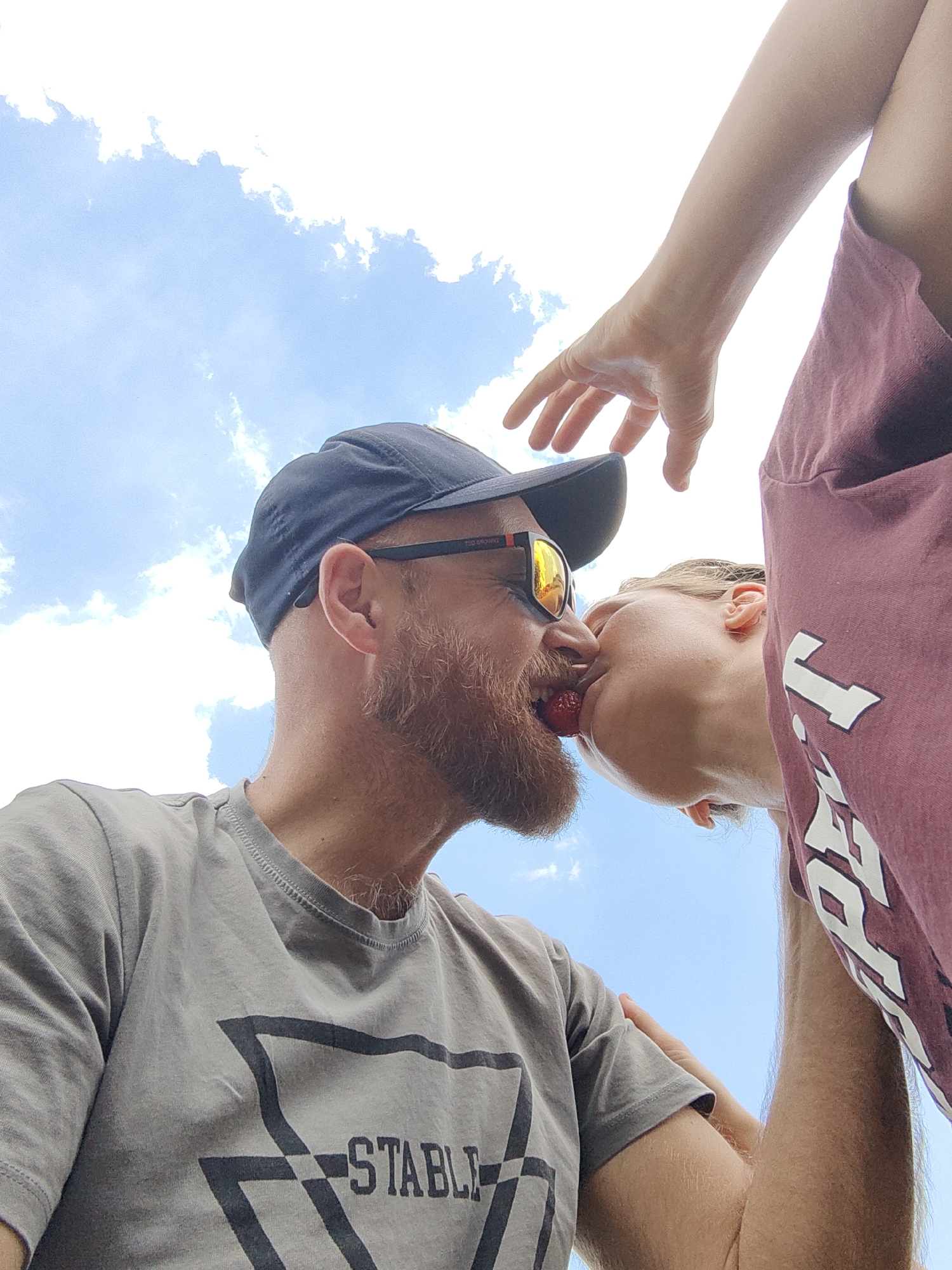 The image shows a low-angle, close-up shot of two people sharing a strawberry from their mouths against a bright blue sky with white clouds. The person on the left, wearing a grey t-shirt and a dark baseball cap, has a beard and is looking towards the right. The person on the right, wearing a maroon t-shirt, is leaning in towards the left person. A hand, presumably belonging to the person on the right, is visible reaching towards the left person. The environment appears to be outdoors, with the sky as the dominant background. The specific location in the city of Зуйское сельское поселение, Ukraine cannot be determined from the image.