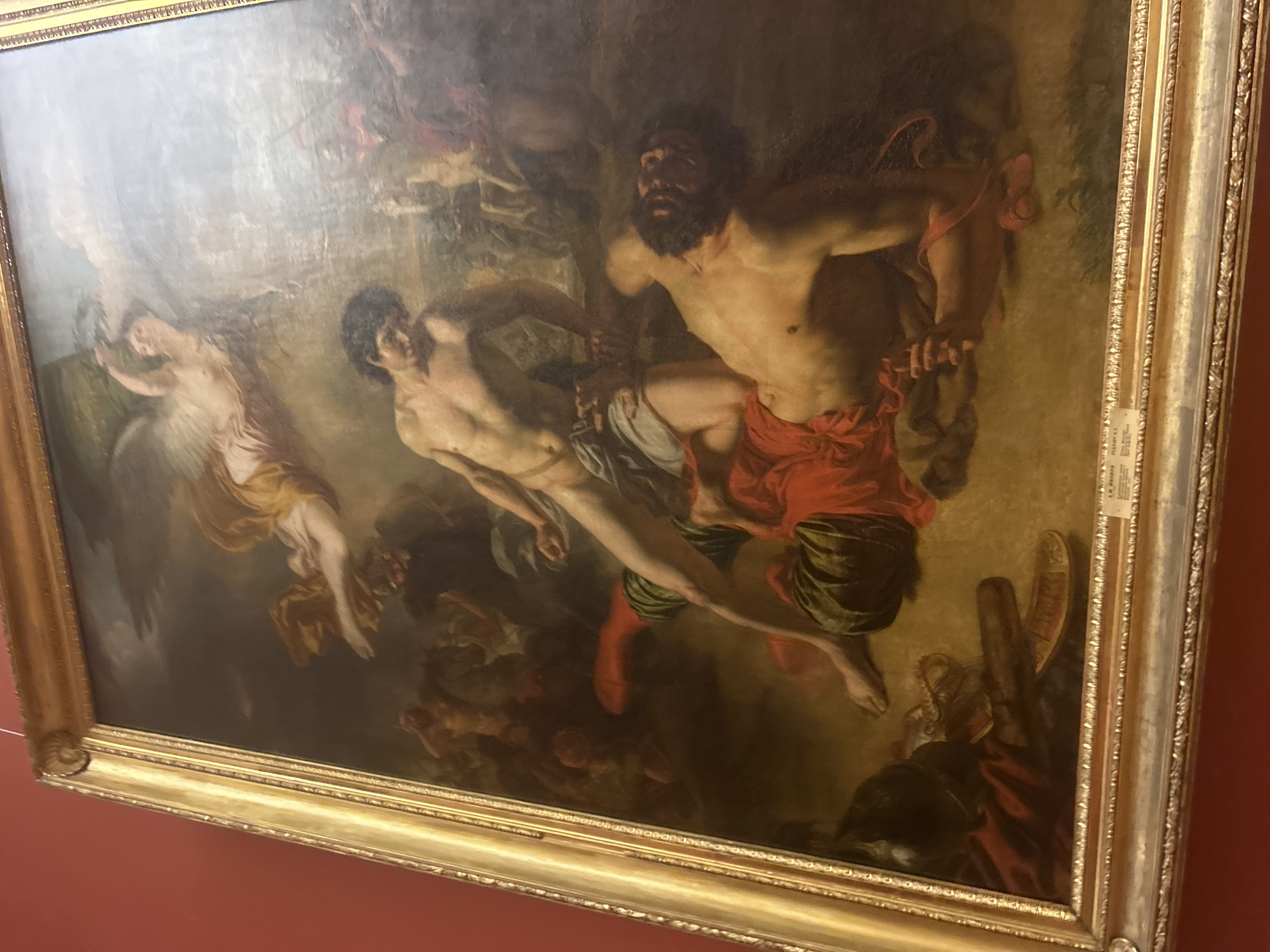 The painting, displayed in a golden ornate frame, depicts a dramatic scene with multiple figures in a battle or struggle. In the foreground, a muscular, pale-skinned man with dark hair is pinning another man to the ground. The man on top is naked from the waist up, wearing a light blue cloth around his hips. The man underneath, with dark, curly hair and a beard, is partially covered by a red garment and green striped fabric. His chest is bare, and he appears to be struggling. Above them, a winged female figure with light hair and a golden sash floats in the sky, reaching down with one hand. In the background, several other figures are visible, some appearing to be engaged in combat, with armor and weapons. The setting is vaguely rocky and earthy, with a dark, cloudy sky. The painting is located in the State Hermitage Museum in Saint Petersburg, Russia, as indicated by the label on the frame which reads "Иванов А. А. (IVANOV A. A.)" and further text that is partially obscured but includes "1863."
