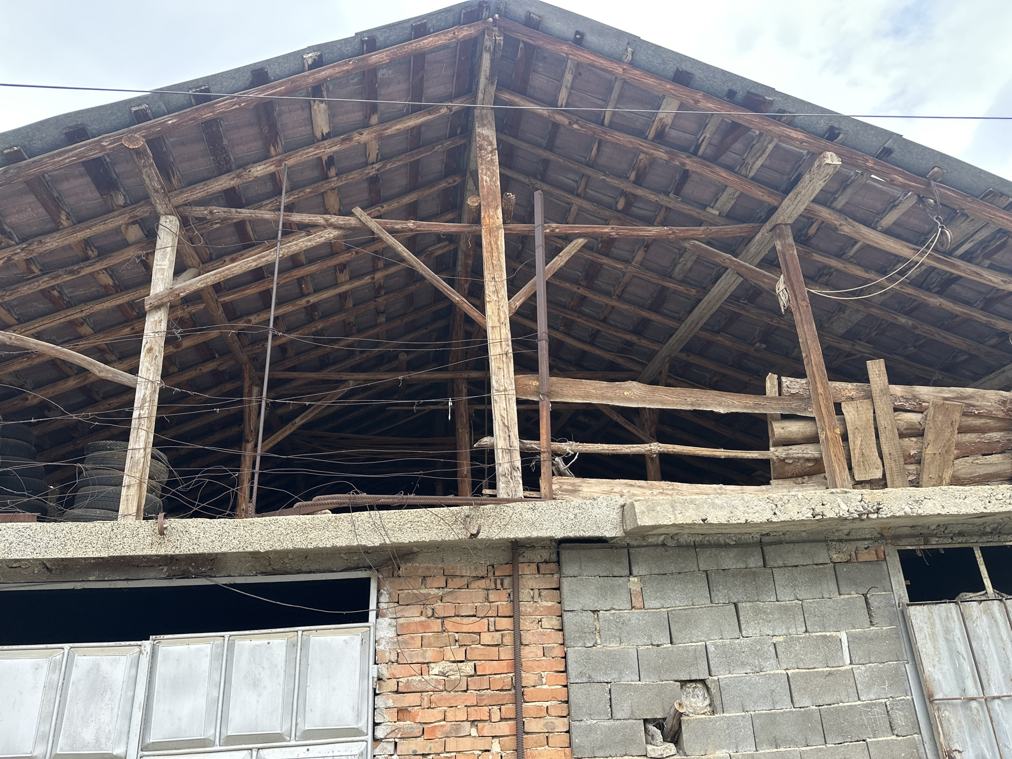 The image depicts a dilapidated structure in Novo Leski, Bulgaria. The lower portion of the building is constructed with a combination of red brick and grey concrete blocks. A large, closed, grey metal gate is visible on the left, and a smaller opening with a partially open metal gate is on the right. Above the brick and concrete wall is a concrete lintel. The upper structure is an open, decaying wooden roof framework, revealing the support beams and roofing structure. There are stacks of tires stored within the building frame. Thin wire runs across the open upper structure. The sky is visible in the background.