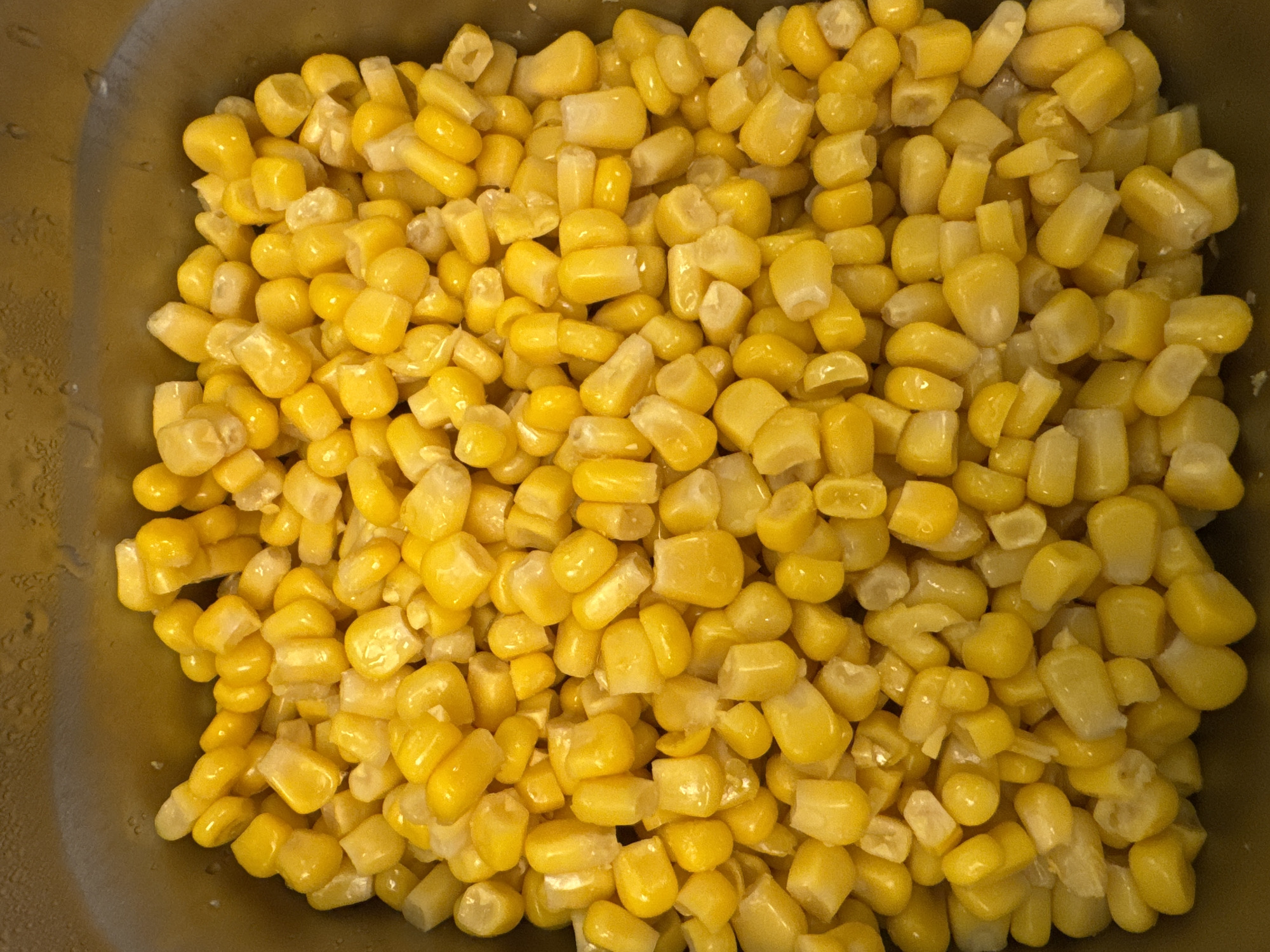 A close-up, overhead shot shows a large quantity of canned corn kernels filling a metallic container. The corn kernels are bright yellow and appear to be cooked and drained. The container has a textured, brushed metal surface, and some condensation or liquid is visible on the inner rim. The scene is static, with no discernible action or interaction. The lighting is even, and the focus is sharp on the corn.