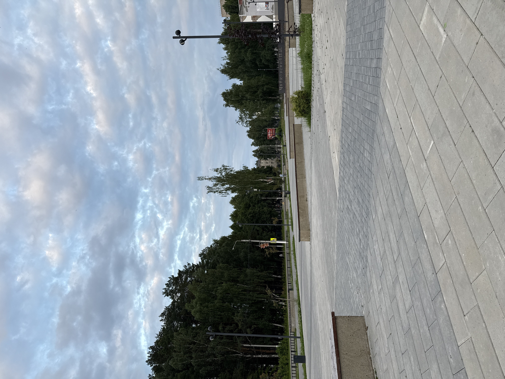 The image depicts an outdoor scene, likely a public park or pedestrian area, in Chaikovsky, Russia. The perspective is from ground level, looking towards a tree-lined avenue. A paved walkway made of grey rectangular stones dominates the right side of the frame, stretching into the distance. To the left, a dense forest of green trees forms a natural barrier.
The sky above is filled with soft, wispy clouds, suggesting it is either early morning or late afternoon. The lighting is diffused, indicating a mild, overcast day. Several streetlights with a minimalist design are visible along the walkway, hinting at an urban or semi-urban setting designed for public use.
In the background, buildings can be seen peeking through the trees, suggesting the presence of a town or city. A prominent signpost with Cyrillic text is visible, partially obscured by trees. The text reads "ЧАЙКОВСКИЙ И.И.", which translates to "Tchaikovsky I.I.", likely a reference to the composer Pyotr Ilyich Tchaikovsky, given the city's name.
There are no people visible in the frame. The overall mood is serene and peaceful, emphasizing the natural beauty of the treeline and the ordered design of the urban infrastructure. The scene appears to be empty and quiet, perhaps before the area becomes busy or after it has cleared.