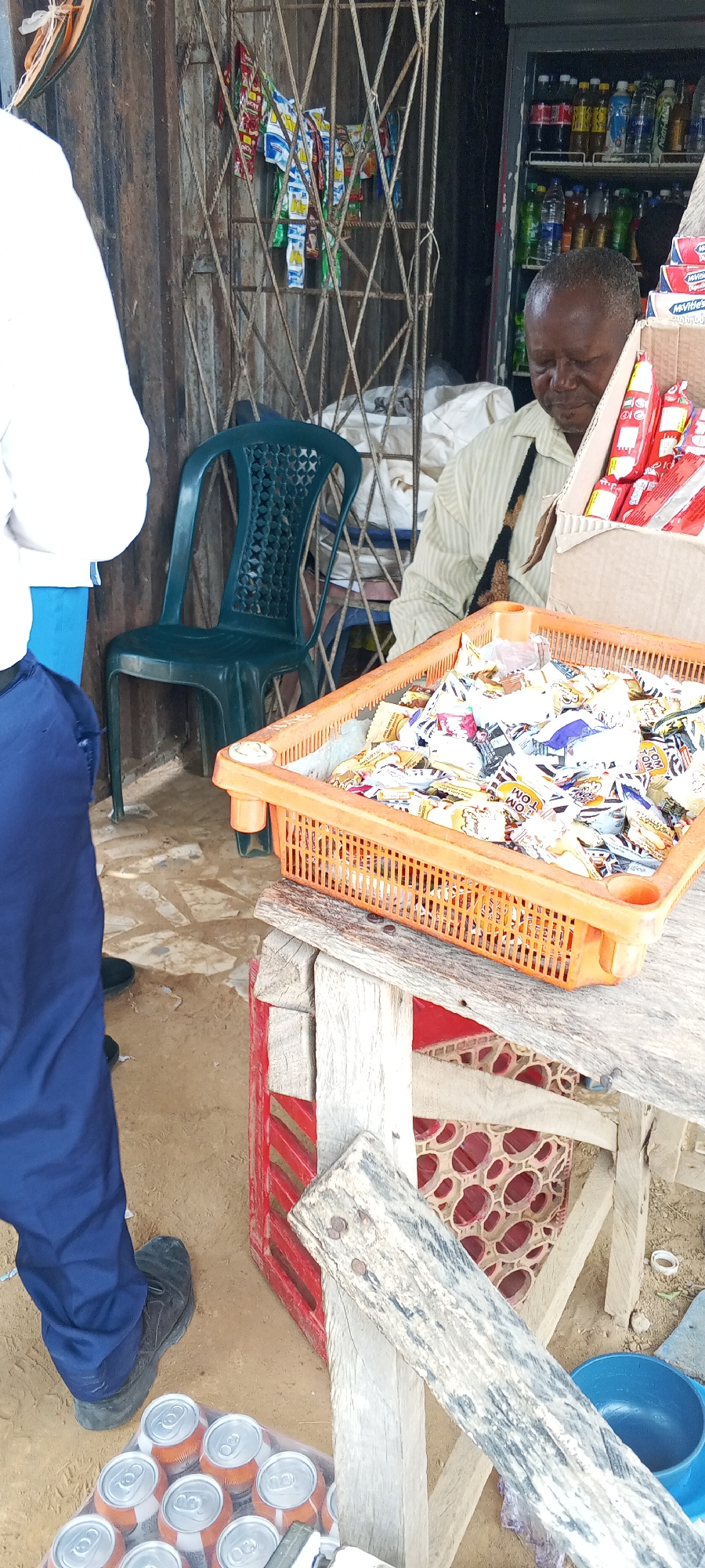 The scene captures a small business operation in Keffi, Nigeria. An elderly man is attending to a street-side shop. He is wearing a light-striped shirt with a dark tie. Before him sits an orange plastic basket filled with small, individually wrapped items, possibly sweets or snacks, sitting on a makeshift wooden table constructed from planks and a red plastic crate. To the right of the table is a blue plastic bucket. Underneath the table, in the bottom left corner, are a dozen cans of a drink, possibly soda.
Behind the man, there's a glass-fronted refrigerator stocked with various beverages. A green plastic chair sits near a wire mesh fence with packets of snacks hanging on it. To the left, a person partially visible, is dressed in a white shirt and blue trousers, possibly a customer. The environment is outdoor, with a sandy ground visible around the setup.