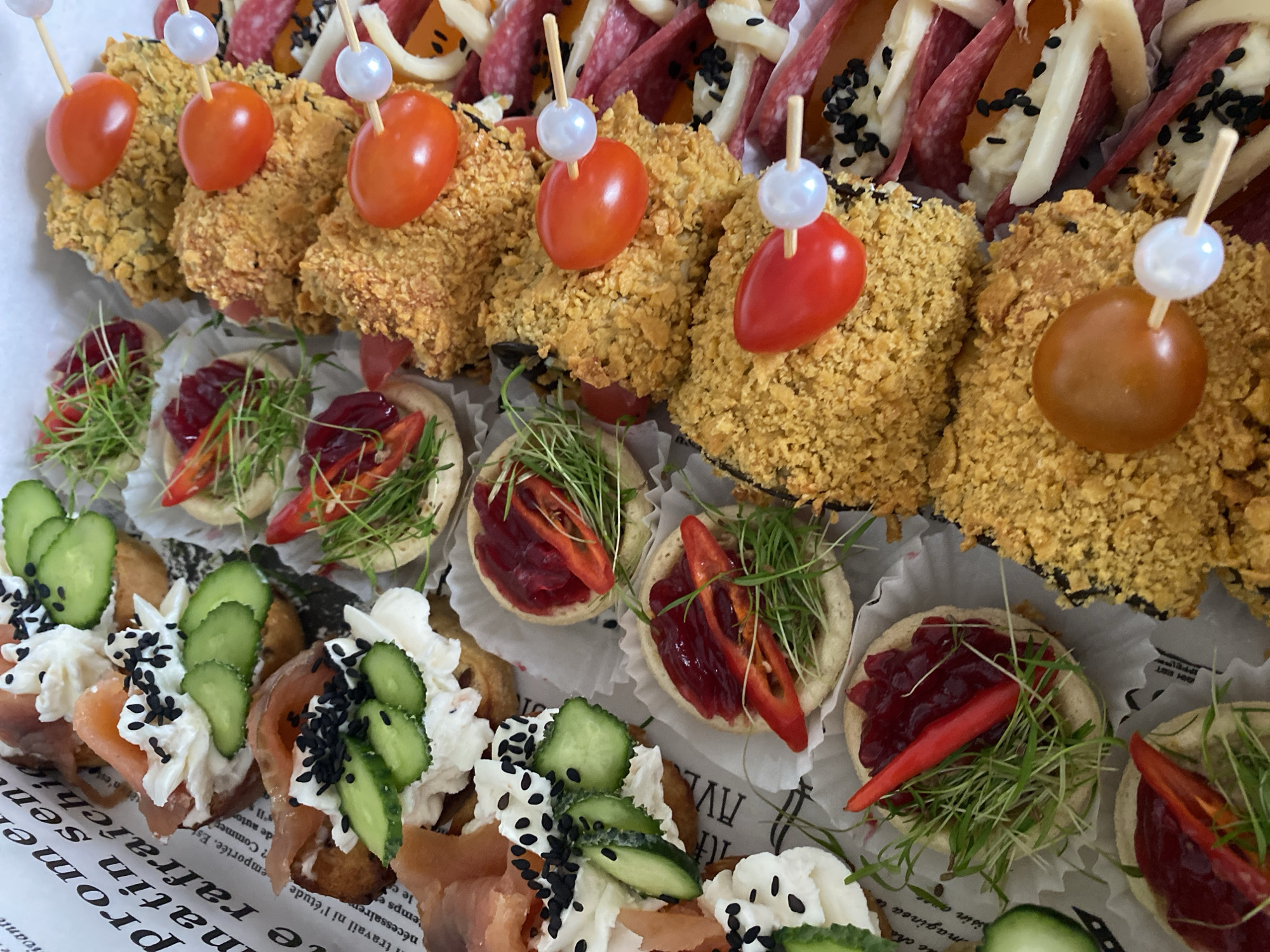 The image displays a close-up overhead view of a platter of assorted appetizers. The appetizers are arranged in neat rows. On the left side of the platter, there are canapés topped with cream cheese, smoked salmon, cucumber slices, and black sesame seeds. Next to these are small circular tartlets filled with a red spread, microgreens, and sliced red chili peppers. In the center and right side of the platter, there are fried, breaded items skewered with cherry tomatoes and small white beads. These are interspersed with folded slices of cured meat and cheese, also dotted with black sesame seeds. The platter itself is set against a backdrop of paper with text, possibly a decorative liner. The environment appears to be an indoor setting, likely a table. No people are visible in the image, and no specific location in Tret'ya Pad', Russia, can be identified.