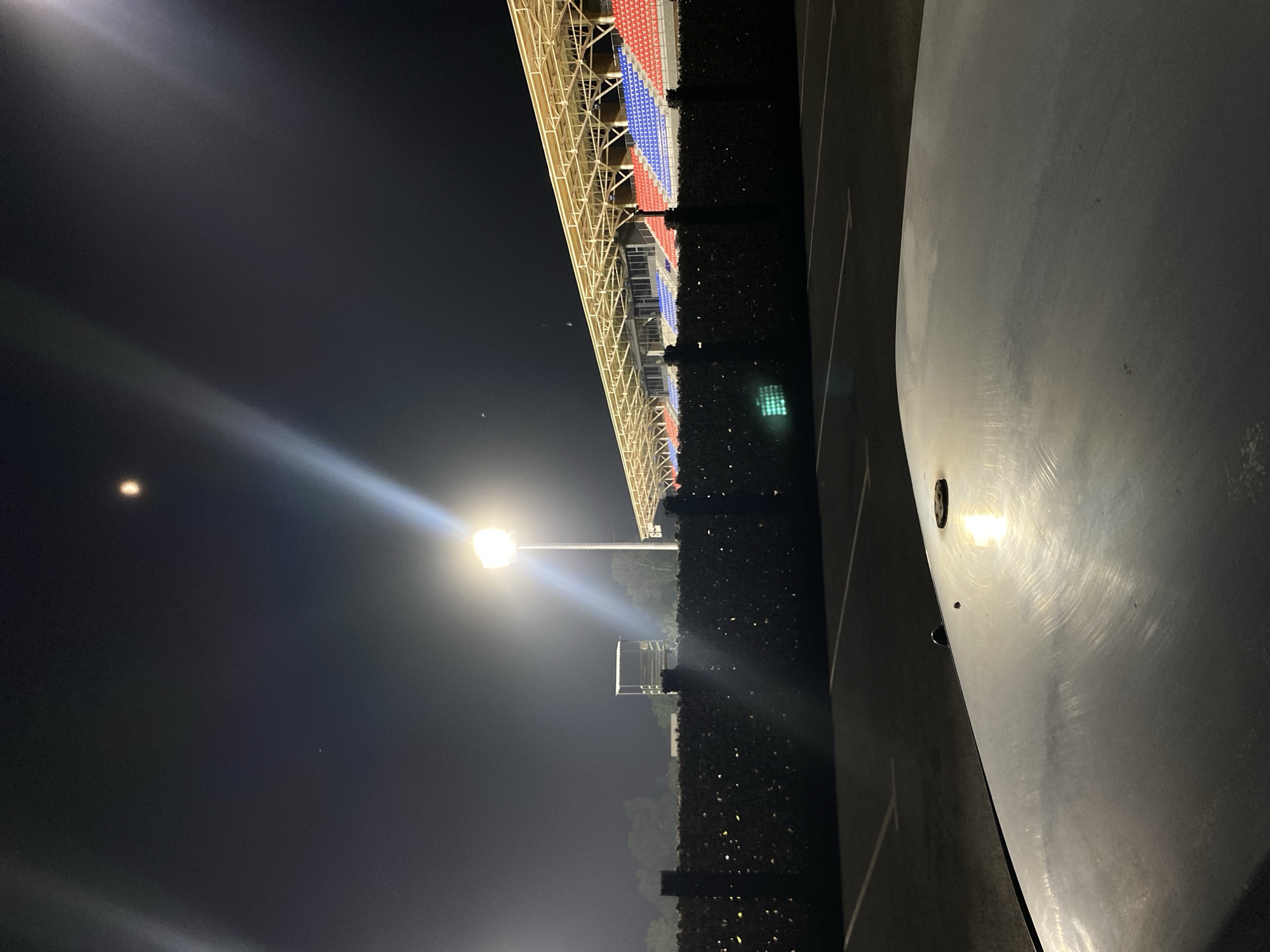 The image depicts a night scene at a stadium in Bishkek City, Kyrgyzstan. A brightly lit stadium floodlight illuminates the dark sky, with a faint moon visible. In the foreground, a car's hood reflects the light. Behind the hood, a dark, textured fence is visible, adorned with numerous small, bright lights that resemble fireflies or LEDs. Beyond the fence, stadium seating sections of red and blue chairs are discernible under a covered grandstand. A tall, empty observation or commentary booth stands to the left of the grandstand. The ground appears to be a dark asphalt parking lot with white painted lines indicating parking spaces.