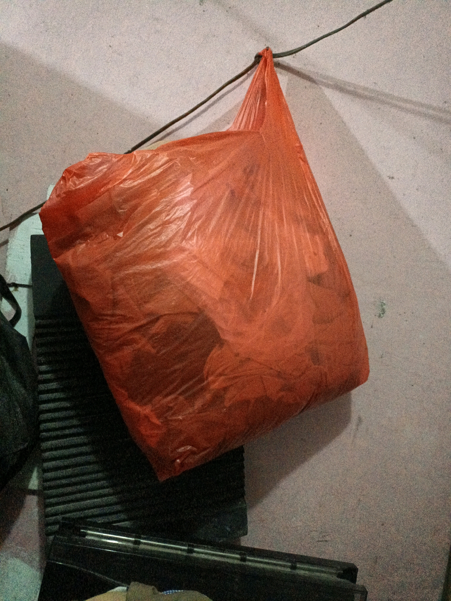 A bright orange plastic bag is hanging from a thin wire attached to a wall. The bag appears full and is casting a shadow on the textured wall behind it. To the left of the bag, a dark, ribbed object is visible, possibly part of some equipment or furniture. Below this, a black electronic device with a metallic stripe runs horizontally across the lower part of the frame. The wall itself is a pale, rough texture with some subtle color variations and small dark specks. The lighting suggests an indoor setting, likely during the day or under artificial light, as indicated by the distinct shadow. The scene is devoid of any people. There is no visible text in the image. The context provided indicates the location is West Jakarta, Indonesia.