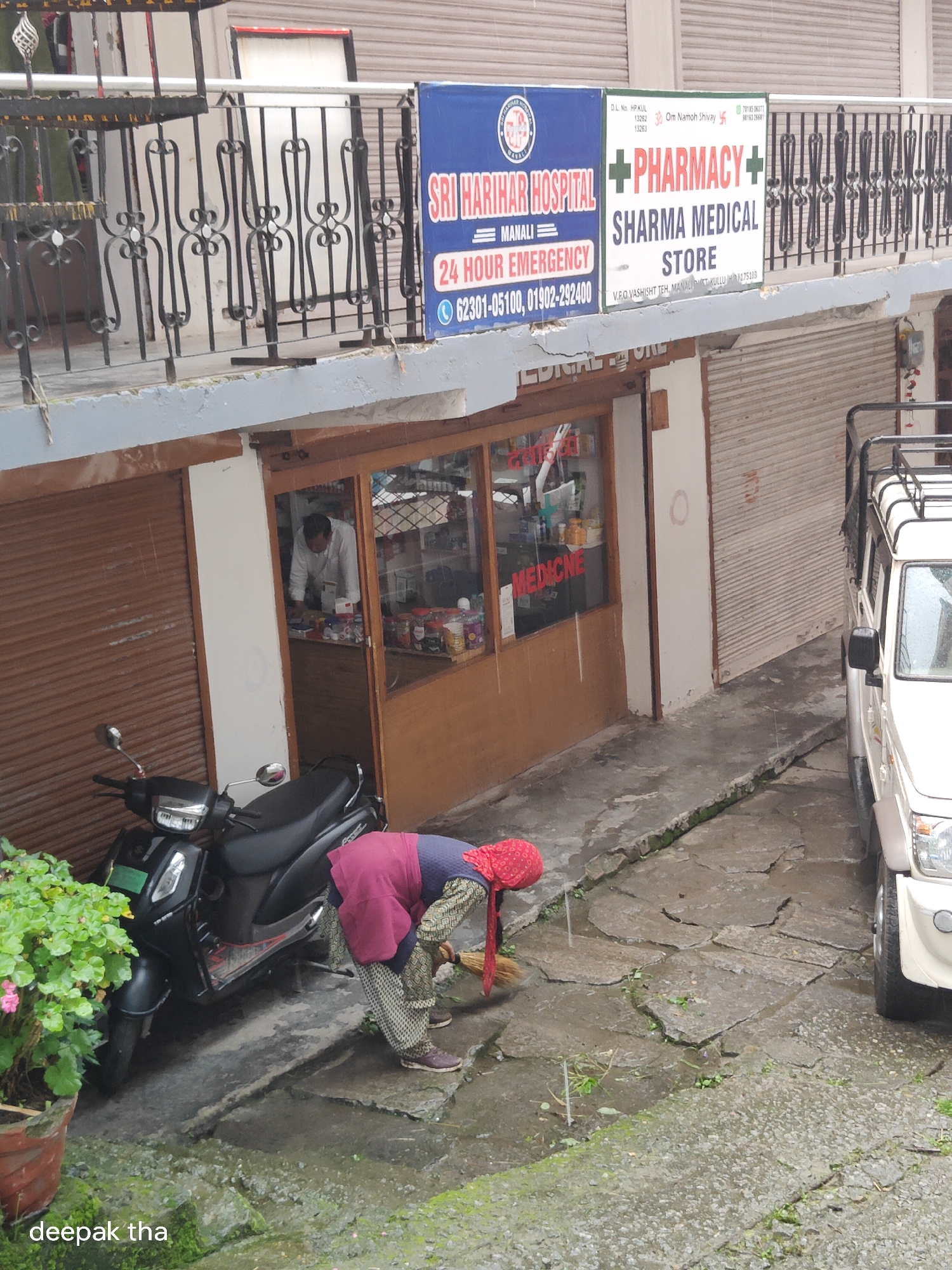 The image captures a street scene in Vashisht, India, featuring a multi-story building with shops and a pharmacy. On the second floor, a sign reads "SRI HARIHAR HOSPITAL MANALI" and another sign indicates "PHARMACY SHARMA MEDICAL STORE." Below, at street level, a pharmacy with a glass storefront displays various products, and a man in a white shirt is visible inside. A black scooter is parked to the left of the pharmacy entrance. In front of the pharmacy, a woman wearing a red headscarf and a patterned skirt is sweeping the wet, uneven stone pavement with a broom. To the right of the pharmacy, a white vehicle, possibly a utility vehicle, is parked. A potted plant with green leaves and pink flowers sits to the far left, and the text "deepak tha" is visible in the lower-left corner. The environment appears to be an outdoor commercial area with wet ground, suggesting recent rain or a damp climate.