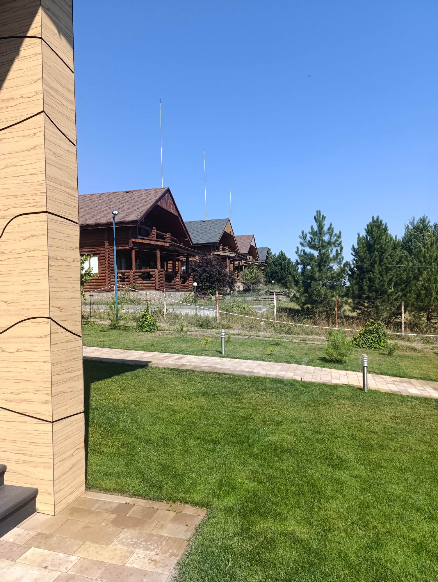 The image shows a sunny day with a clear blue sky over a rural landscape. In the foreground, a modern building with a light-colored, textured facade stands on the left. A paved walkway made of tan square and rectangular stones leads through a well-maintained green lawn.
In the middle ground, a line of wooden log cabins with brown roofs are nestled on a gentle slope. These cabins appear to be in a resort or residential area, possibly in the village of Bulatselivka, Ukraine. Some of the cabins have porches with wooden railings and visible windows. A few trees, including tall pine trees, are situated behind the cabins, forming a natural backdrop. There is a wire fence running along the property line in front of the trees. Several modern, cylindrical path lights are visible along the walkway. The overall scene suggests a peaceful, outdoor setting with a blend of modern architecture and natural elements.