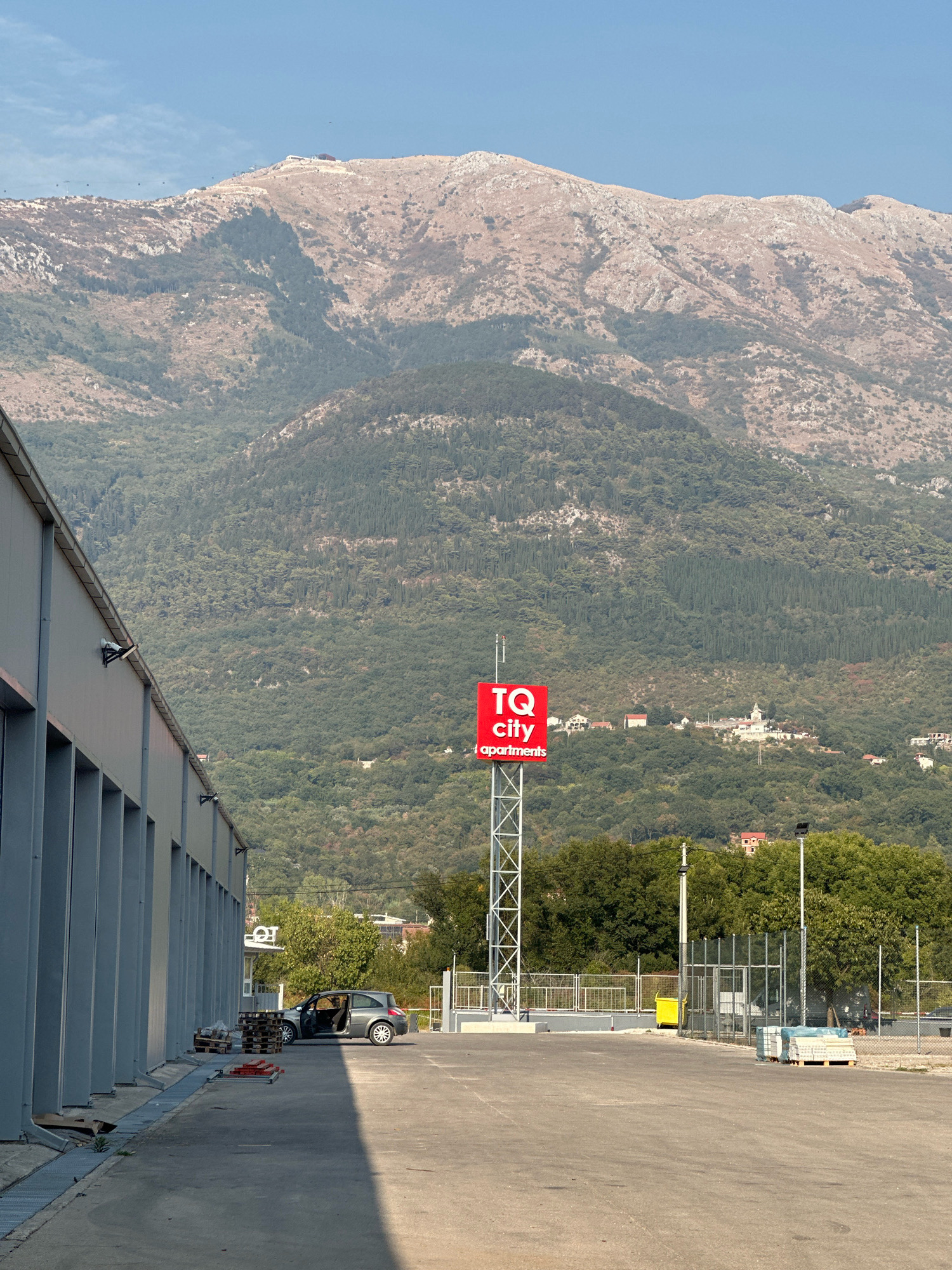 In Radanovići, Montenegro, a gray sedan is parked with its driver's side door open in front of a long, gray industrial building. Adjacent to the car are several wooden pallets, some empty and others stacked with items. Further into the scene, a tall red sign with white text reading "TQ city | apartments" stands on a metal tower. The area is paved, with a chain-link fence visible in the background, beyond which are trees and a village with white buildings nestled on a hillside. Looming behind the village and trees is a large, rugged mountain range with sparse vegetation on its slopes. The sky above is clear and blue.