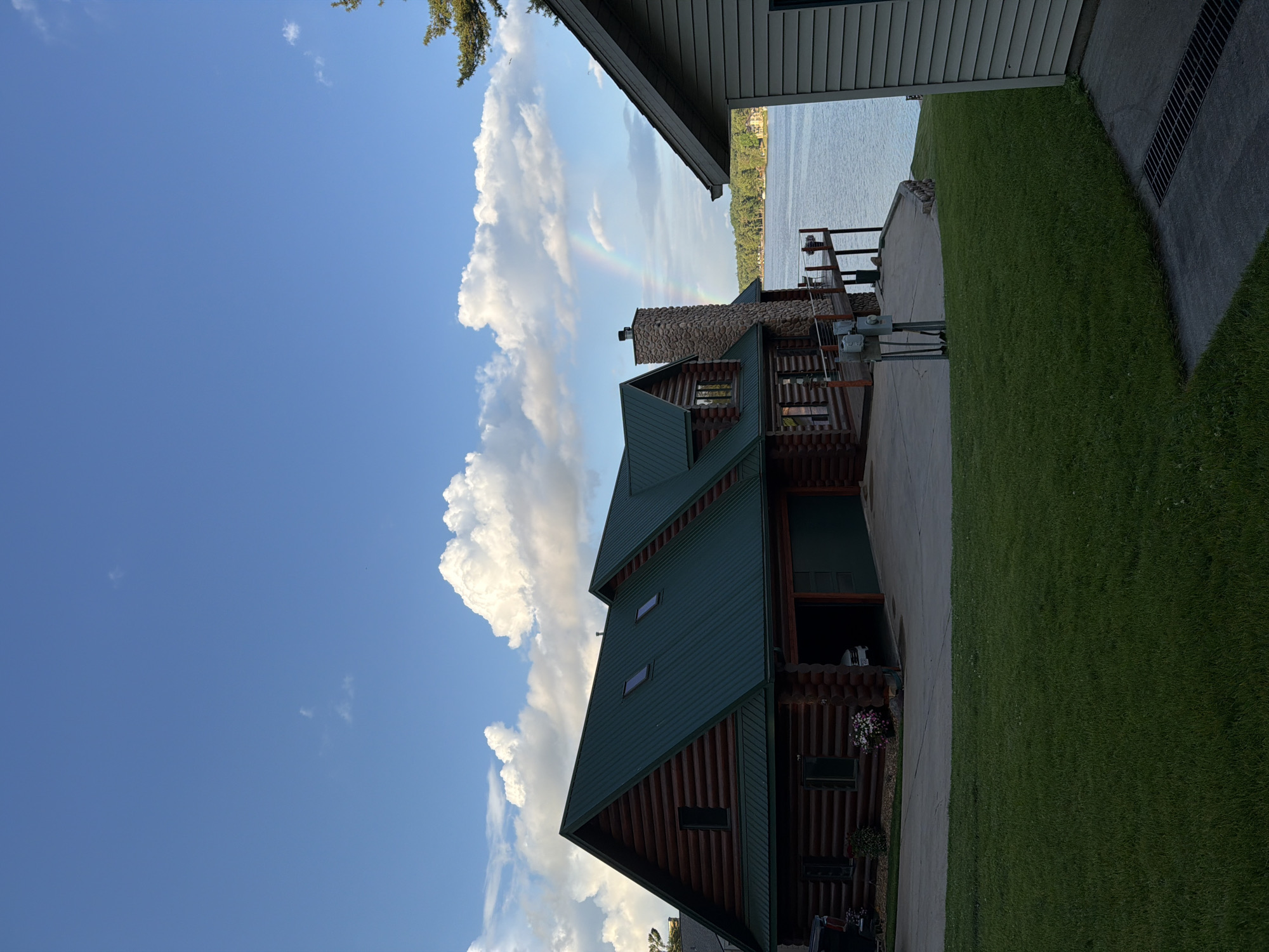 The image captures a detailed outdoor scene of a lakeside residential property in Lake Delton, United States of America. The primary subject is a large, two-story log cabin-style house positioned on the left, featuring dark brown log construction and a dark green corrugated metal roof with multiple gables and two skylights. A two-car garage with dark green doors is integrated into the front of the house; one door is open, revealing a white vehicle inside. A dark truck is partially visible parked on the far left. A large, multi-toned stone chimney ascends from the right side of the house, adjacent to a wooden deck equipped with metal railings that extends towards the lake. Below the deck, electrical utility boxes and conduit are visible.
In the foreground, a well-maintained green lawn occupies the lower half of the image, bordered by concrete driveways and walkways. A concrete path with a metal grated drain is present in the extreme lower right. A partial view of another building, clad in light-colored horizontal siding with a dark roof edge, is visible on the far right.
The house is situated on the shore of a wide, calm lake, which dominates the middle-ground. A faint rainbow arcs across the sky over the lake, to the right of the main house. The far side of the lake reveals a densely wooded shoreline with scattered residential structures. The sky above is clear blue with several scattered white cumulus clouds, particularly concentrated above the main house and extending towards the center-right. A single pine tree branch is visible in the upper right corner of the frame. No individuals are present in the image. The lighting suggests daytime conditions.