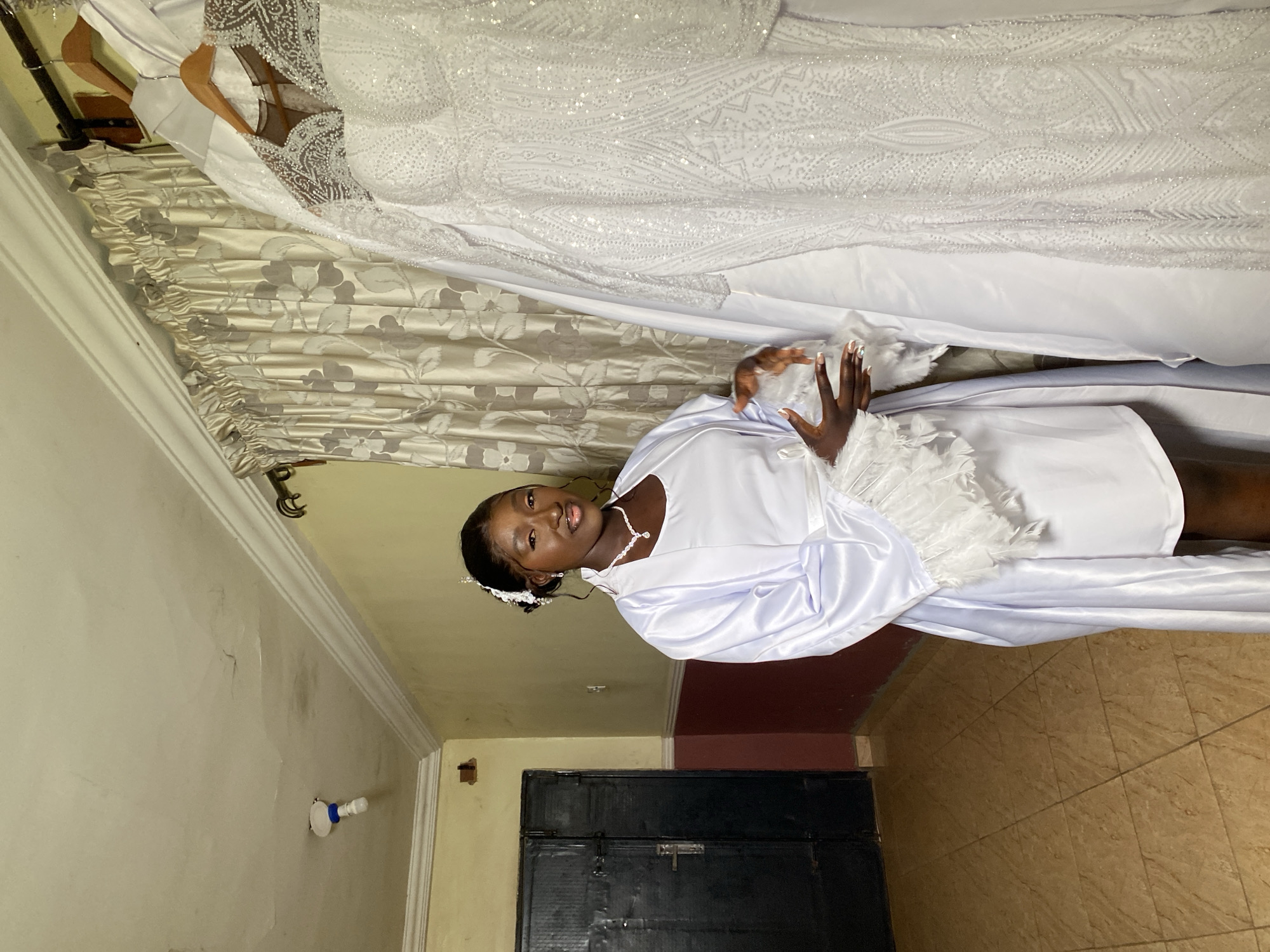 A medium shot of a woman with dark hair, wearing a white robe with feather details around the shoulders and a white necklace, posing indoors. Above her is a wedding dress hanging on a hanger against a patterned curtain. The location appears to be a room within a building in Barikin Sabon Gidan Kanat, Nigeria, judging by the type of flooring and door present. The walls are painted a light color, with some discoloration. A light fixture is visible on the wall. A black door is to the left.