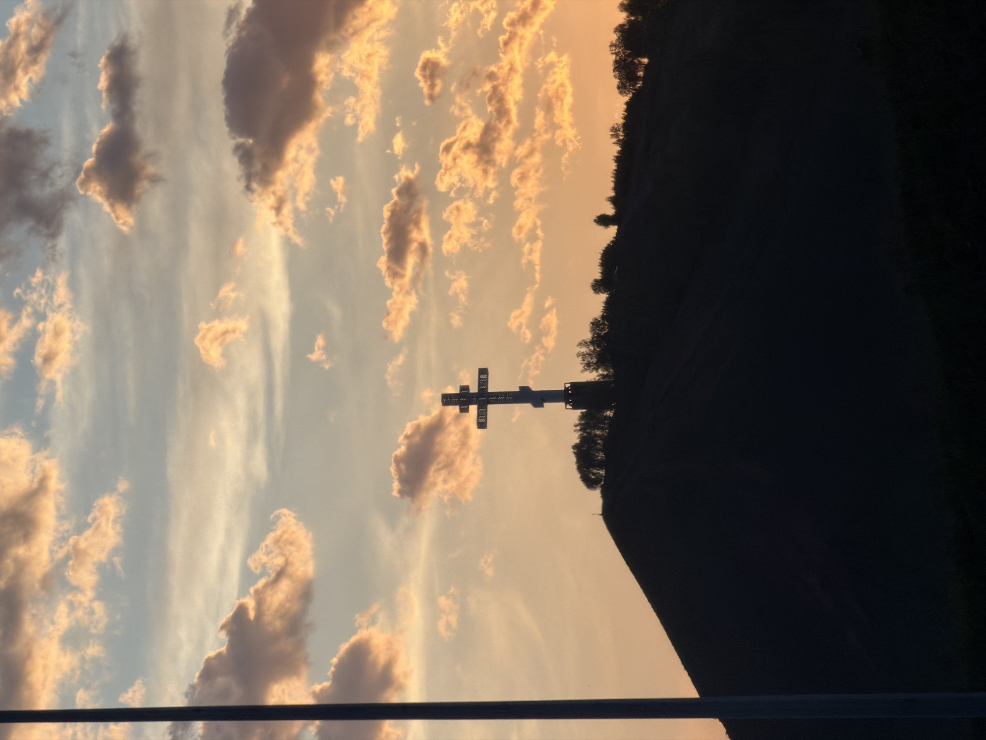 The image captures a sunset view in Krasnoyarsk, Russia, dominated by the silhouette of a large Orthodox cross atop Karaulnaya Mountain. The sky displays a gradient of colors, with hues of orange and yellow near the horizon transitioning to light blue higher up. Scattered clouds are present, illuminated by the setting sun. The dark silhouette of the mountain is to the right of the cross. An object near the bottom of the image is out of focus.
