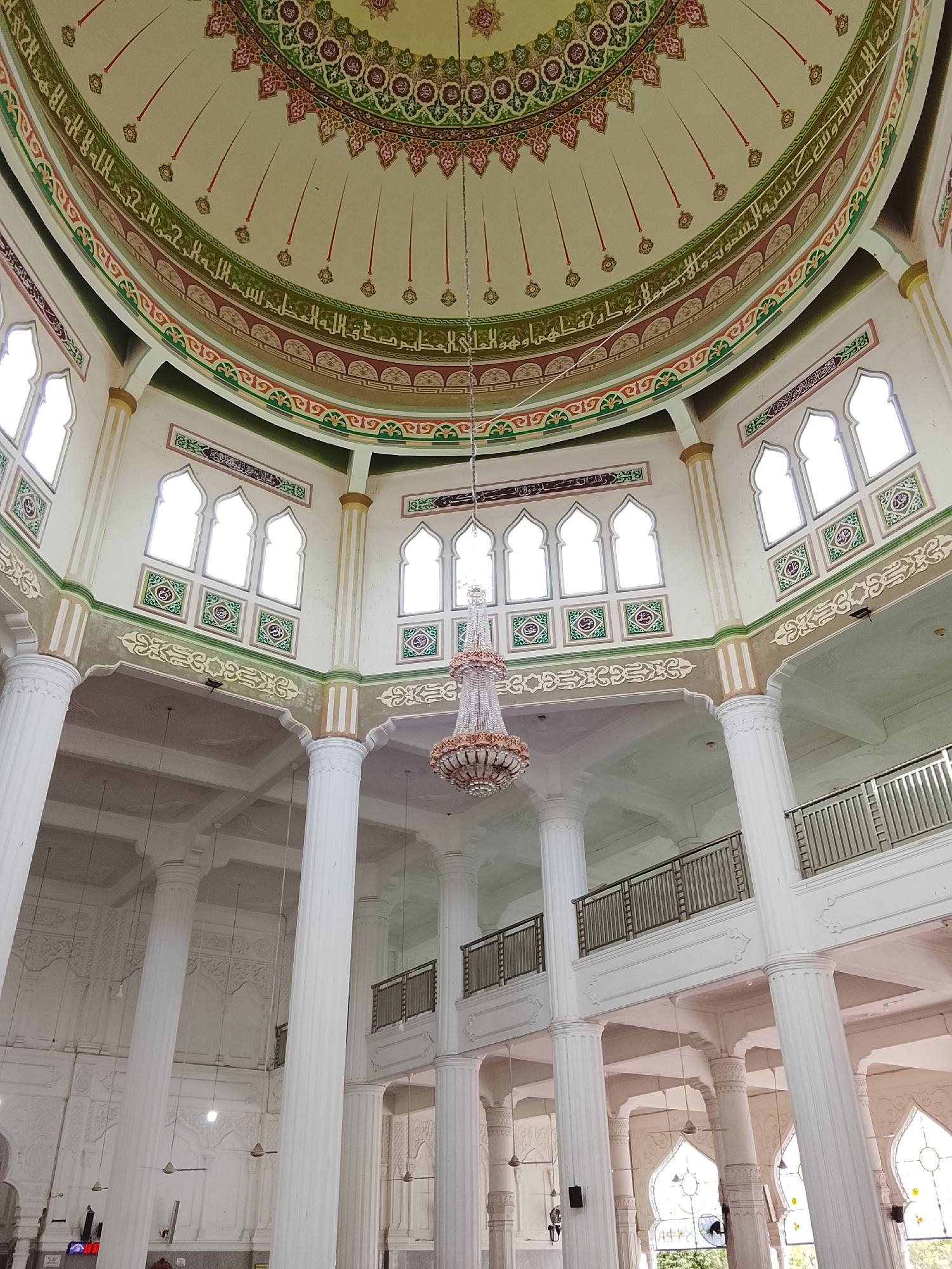 The image shows the interior of the Islamic Center Mosque in Lhokseumawe, Indonesia. The camera is positioned looking upwards towards the domed ceiling, which is adorned with intricate Arabic calligraphy and colorful geometric patterns in shades of green, red, and gold. Below the dome, large white pillars support the structure, and a grand chandelier hangs from the ceiling. The walls feature rows of arched windows with decorative panels below them. The overall impression is one of grandeur and religious devotion. The mosque is a prominent landmark in Lhokseumawe, a city located on the northern tip of Sumatra island, Indonesia.