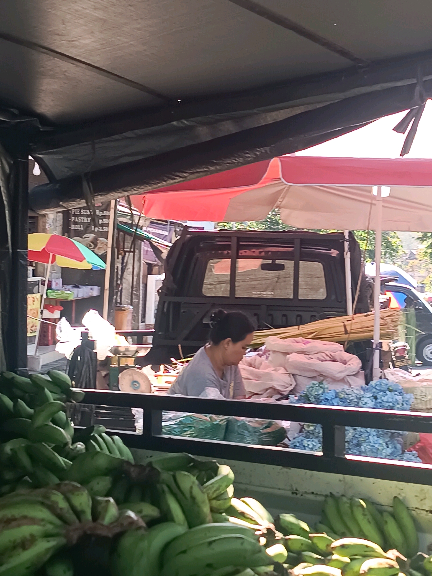 This image depicts an outdoor market scene in Tabanan, Indonesia. In the foreground, a large quantity of green bananas is piled in a crate. Behind the bananas, a woman wearing a grey t-shirt is visible, appearing to be engaged in market activities. She is positioned behind a metal railing. Beyond the railing, several large sacks, possibly filled with goods, are stacked. Further in the background, a black pickup truck is parked, and an assortment of items, including what appear to be bundles of dried grass or reeds, are loaded in its bed. Two umbrellas, one red and white and another red, are open, providing shade. In the mid-ground, a smaller multi-colored umbrella stands next to a stall with various items on shelves. The overall environment suggests a bustling marketplace under a covered structure, with a hint of sunlight filtering through the canopy.