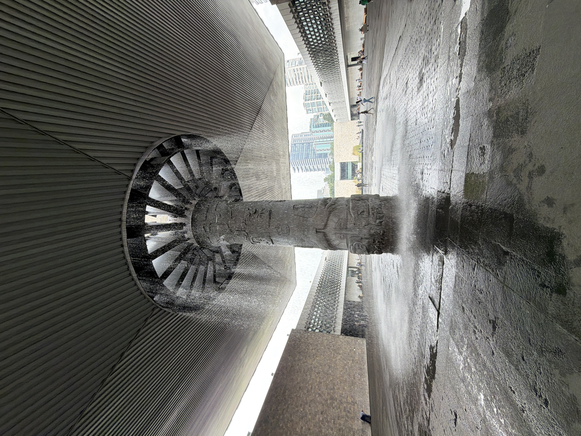 The image captures a low-angle view of the water feature at the Museo Nacional de Antropología in Mexico City. A large, carved stone column, possibly a replica of an ancient artifact, stands in the foreground, with water cascading from a circular structure above it, creating a mist that partially obscures the scene. The column is intricately decorated with relief carvings.
The water falls onto a dark, wet stone courtyard, with reflections visible on the wet surfaces. The courtyard is partially covered by a large, ribbed, metallic canopy overhead, which extends outwards, creating a sheltered area. In the background, the distinctive architecture of the museum buildings is visible, characterized by textured stone walls and modern design elements with patterned facades.
Several people are present in the mid-ground and background, casually walking or standing in the courtyard. Some appear to be observing the water feature or interacting with the surroundings. The environment suggests an overcast or slightly rainy day, with the water feature appearing to be actively operating. The wider cityscape with tall buildings is visible in the distance, indicating an urban setting. A lone figure is crouched near the left wall of the museum building.