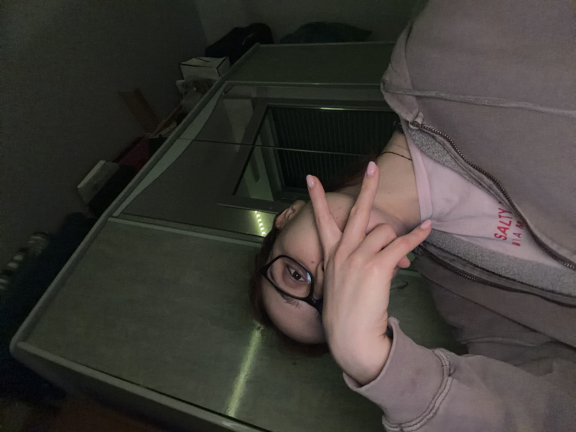 This is a medium shot, high-angle, selfie-style photo of a person with red hair and glasses. The person is wearing a light purple zip-up hoodie over a pink t-shirt that has "SALTY MIAMI" printed on it in red text. The person is holding their right hand up in front of their face, with their index finger and thumb extended in a gesture. The background appears to be an interior space with a large, dark-colored wardrobe or closet with mirrored doors. The lighting is dim, with some string lights visible on the left side of the frame, suggesting an indoor, possibly residential, setting. There is no discernible connection to the city of Strasbourg, France.