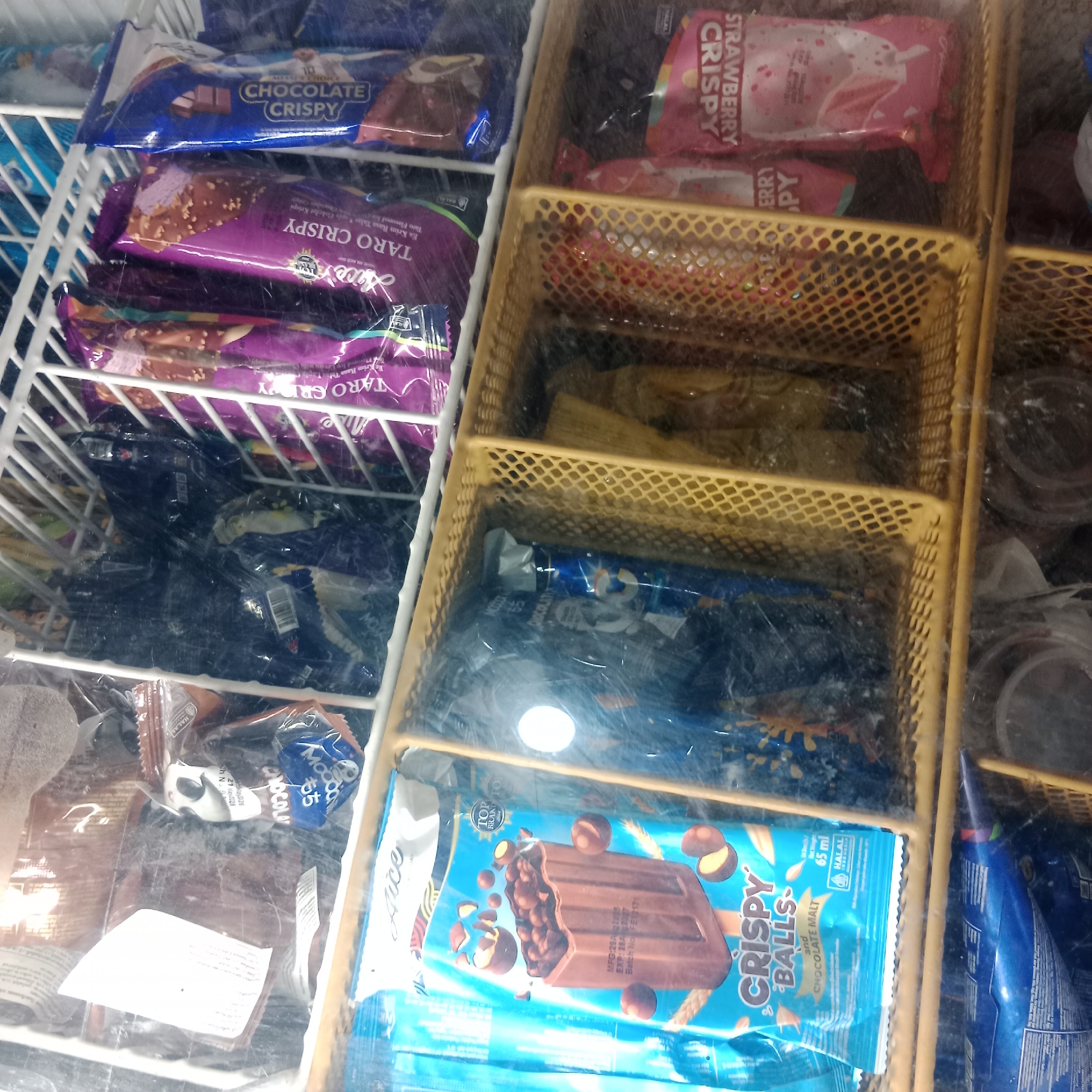 This image shows the interior of a freezer chest, likely at a shop or minimarket in Tegowanu Wetan, Indonesia. The freezer is filled with various packaged frozen desserts, predominantly ice cream bars. On the left side, there are rows of ice cream bars with purple packaging labeled "TARO CRISPY" and blue packaging labeled "CHOCOLATE CRISPY". To the right, a yellow wire basket contains several pink packages labeled "STRAWBERRY CRISPY". Below this, another yellow wire basket holds assorted items, including what appear to be smaller packaged desserts. Further down, more ice cream bars are visible, with one prominent blue package displaying a chocolate bar covered in crispy balls, labeled "CRISPY BALLS and CHOCOLATE MALT". The overall environment appears to be a commercial setting due to the organized display of products in a freezer.