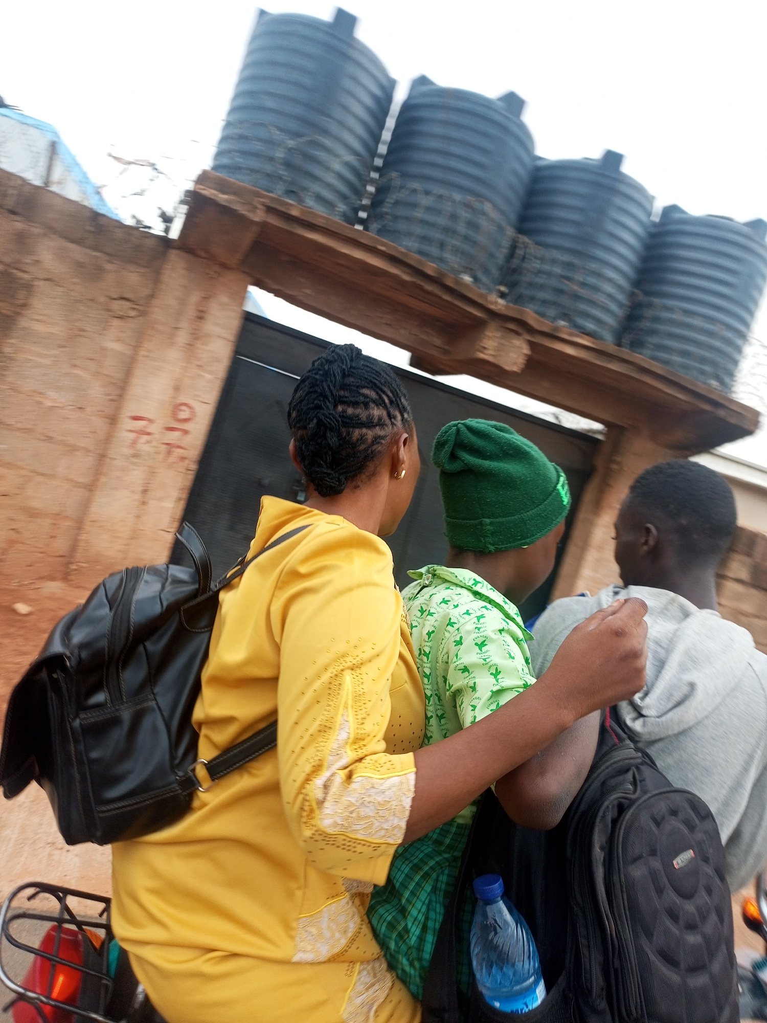 A medium shot captures three individuals on a motorcycle in Tempure, Nigeria. A woman in a yellow outfit and black backpack drives, while two passengers ride behind her: a person in a green hat and patterned shirt, and another in a grey hoodie with a black backpack. A water bottle is visible in the side pocket of one of the backpacks. Above them, four dark grey water storage tanks are stacked on a wooden platform affixed to a light brown wall. A dark metal gate is visible in the background behind the platform. The sky is overcast.