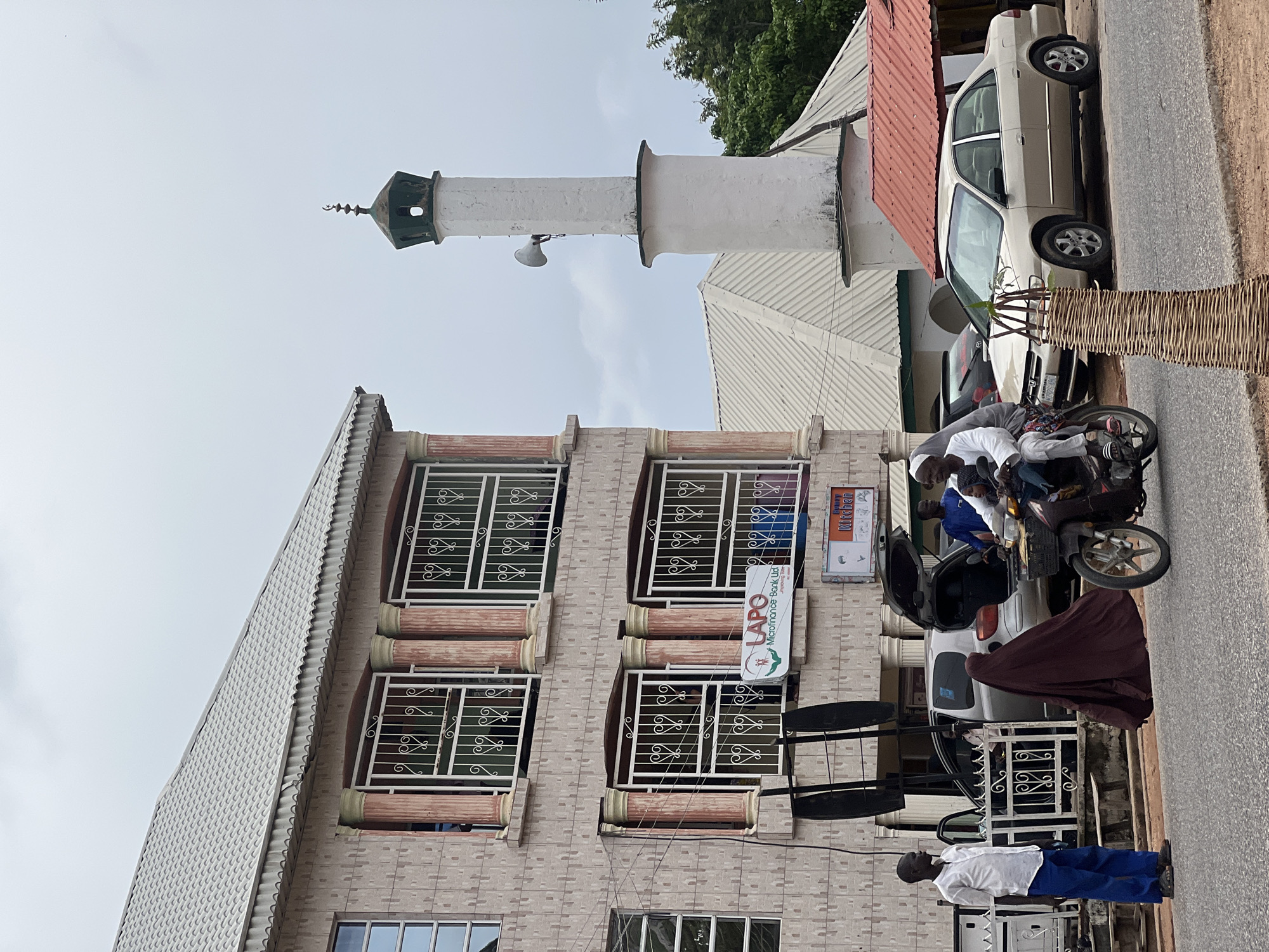 In the city of Jalingo, Nigeria, a street scene unfolds with a multi-story building on the left and a mosque minaret to the right. The building, constructed with pale pinkish-tan bricks, features decorative columns around its windows, which are fitted with white metal grilles. A sign for "LAPO Microfinance Bank Ltd." is visible on the building's facade.
In the foreground, a man in a white shirt and blue trousers stands on the sidewalk. A motorcycle with two occupants, one in a white hijab and the other wearing white clothing, rides on the street. A woman in a dark maroon hijab walks near the motorcycle. A silver car with its trunk open is parked on the right side of the street, with a man standing nearby. Further to the right, a light-colored sedan is parked. The sky is overcast, suggesting a cloudy day. The environment appears to be an urban street with paved roads and sidewalks.