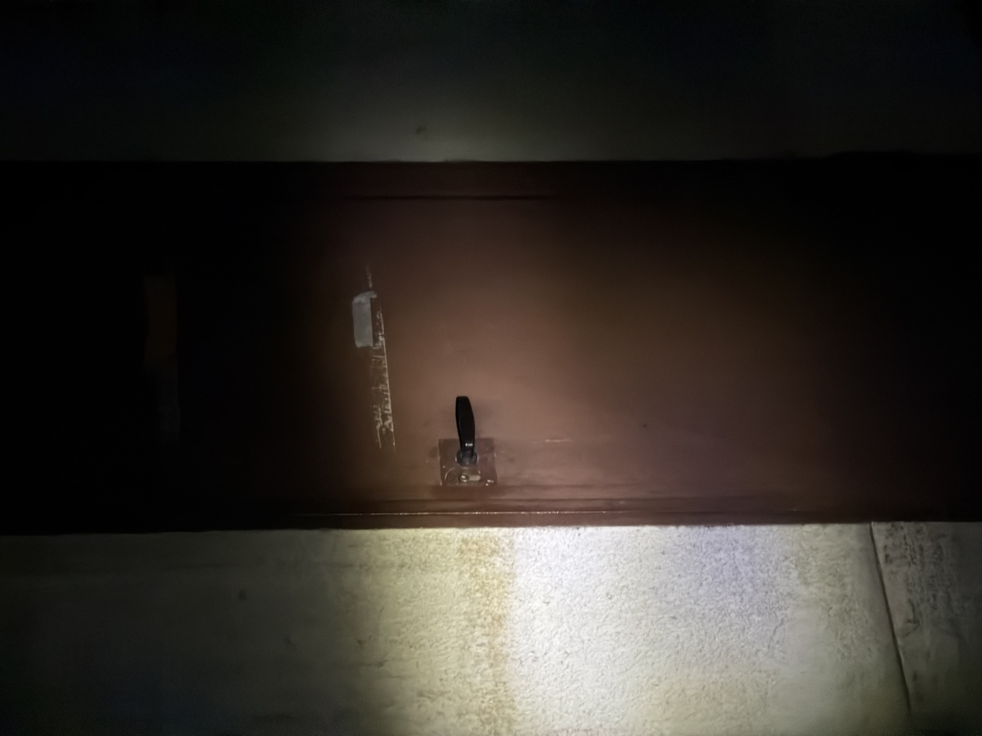 A low-angle, indoor shot depicts a dark, possibly wooden, cabinet against a dimly lit background. Mounted on the cabinet's surface are two metal fixtures: one on the left, and a switch mechanism with a black handle positioned in the center. The lower part of the image reveals a textured surface, possibly a rug or carpet, illuminated by an unseen light source. The location of the image is Unknown, Unknown. There are no people visible in the image.