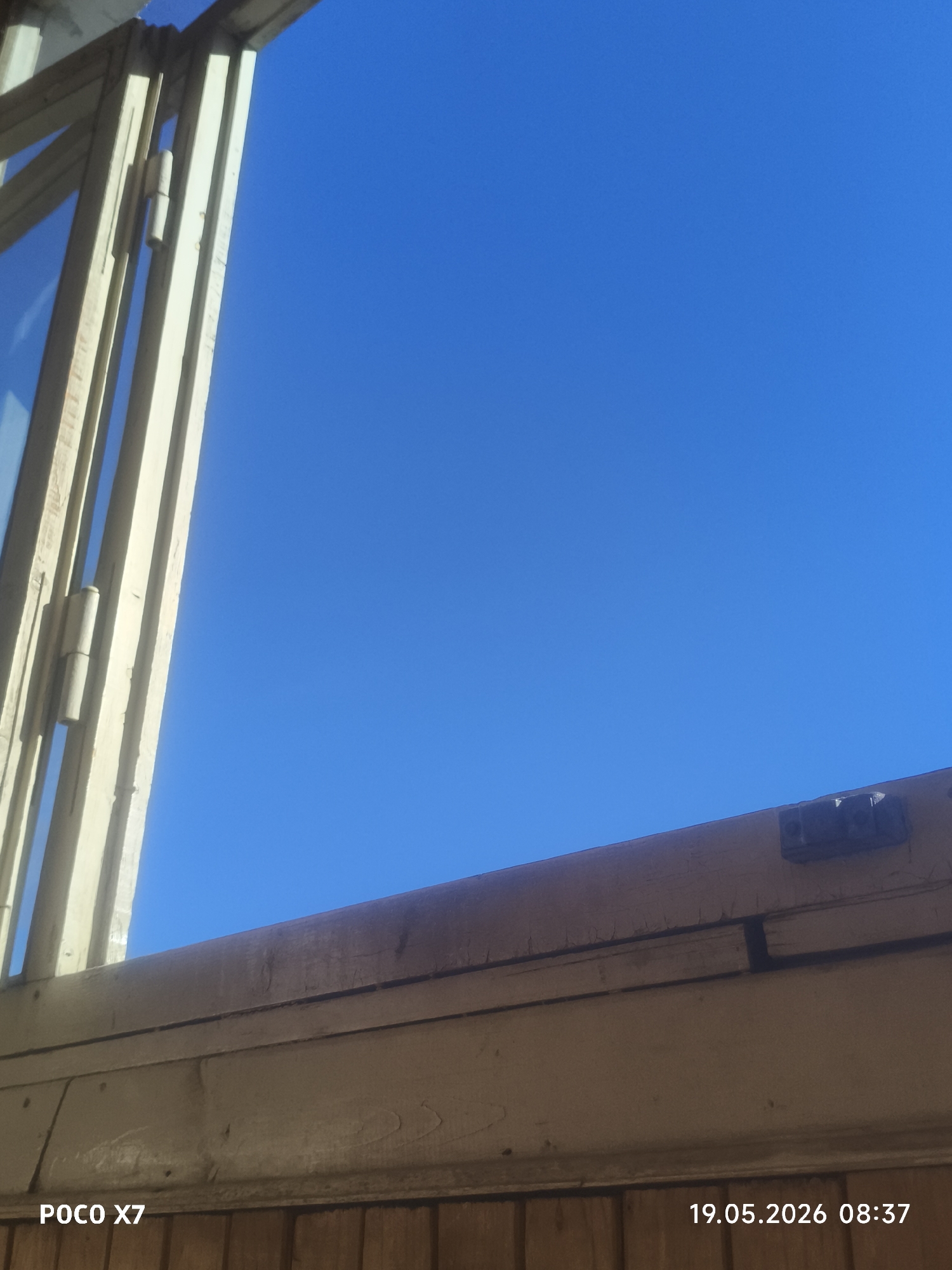 A low-angle shot presents a view through an open window frame, revealing a clear blue sky. The window frame is constructed of light-colored wood, and hinges are visible on the left side, indicating the window's opening mechanism. A portion of the window sill and the wall beneath it are visible, made of wood with a horizontal plank pattern. The camera's timestamp, "19.05.2026 08:37," and the device model, "POCO X7," are displayed in the lower corners of the image. The location is Unknown, Unknown.