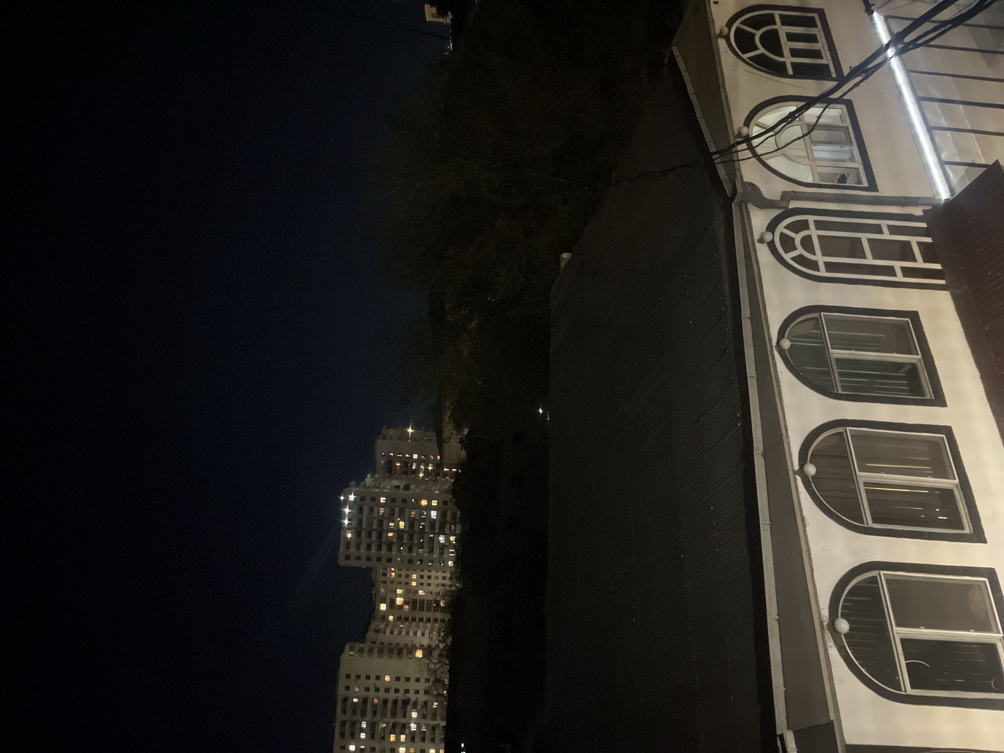 The image depicts a nighttime urban scene, likely taken in Yerevan, Armenia. The scene is divided between darkness and light.
On the left, a tall, illuminated skyscraper dominates the view against a dark sky. The building features numerous windows glowing with light, suggesting residential or office occupancy.
On the right, a building with white facade is visible. The architecture features a series of arched windows with white frames and dark interiors. A dark wall or structure is partially visible next to the building with arched windows, casting a shadow and contrasting with the white facade.
In the middle ground, dark foliage is seen, providing a natural barrier between the two architectural structures. There are also electrical cables visible in the middle ground.