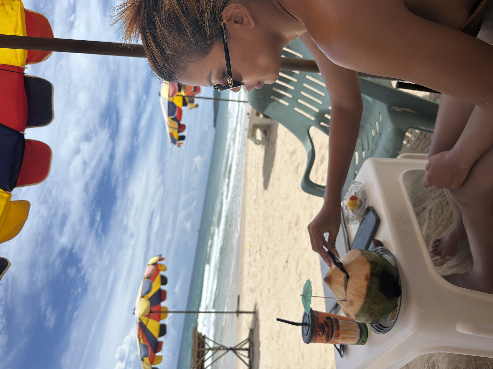 A person is seated at a white plastic table on a sandy beach in Choeng Thale, Thailand, partially shaded by a colorful umbrella. The person, facing right in profile, wears sunglasses and has their hair pulled up. They are reaching for a halved coconut with a straw inserted, which sits on a blue and white plate. A cup with a beverage and a small paper umbrella is also on the table, along with a mobile phone and a small dish containing fruit. In the background, the ocean waves crash onto the shore under a cloudy sky. Two other beach umbrellas are visible further down the beach, along with a wooden structure. A green plastic beach chair is behind the table.