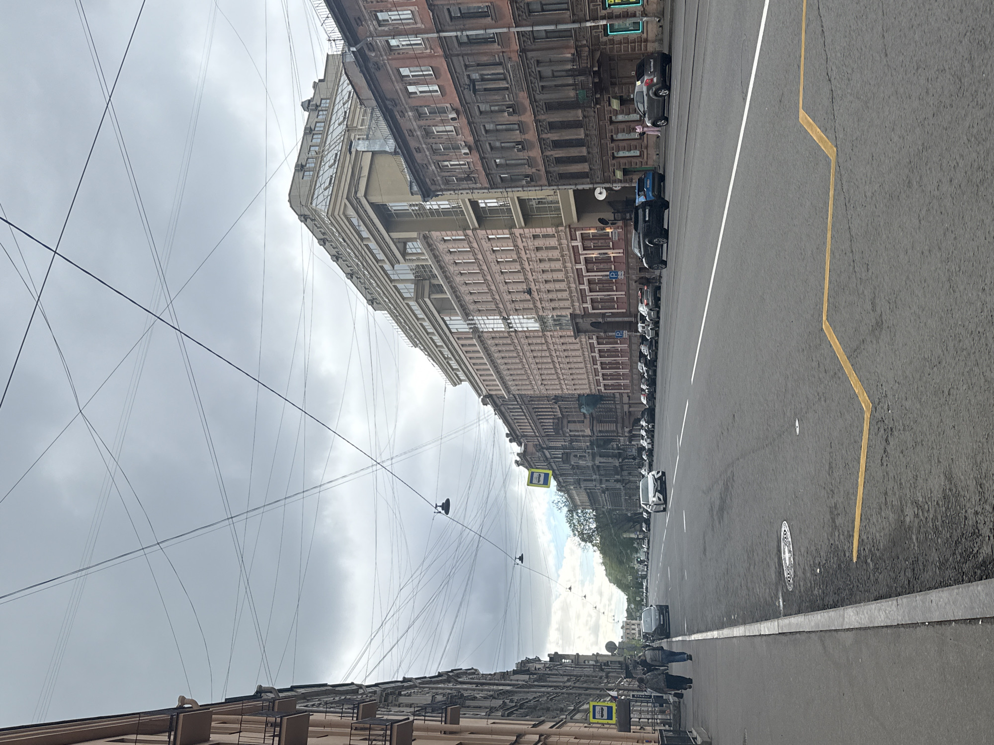 This image depicts a street scene in Saint Petersburg, Russia. The street is lined with buildings that are several stories high and appear to be older architectural styles. There are overhead wires crisscrossing the sky.
On the right side of the street, cars are parked, and there is a lane with a painted yellow zigzag line. A pedestrian sidewalk is visible on the lower right side where people are walking.
The sky is overcast. In the distance, trees and buildings are visible, suggesting the street extends quite far. Pedestrian crossing signs are visible on the side of the street.