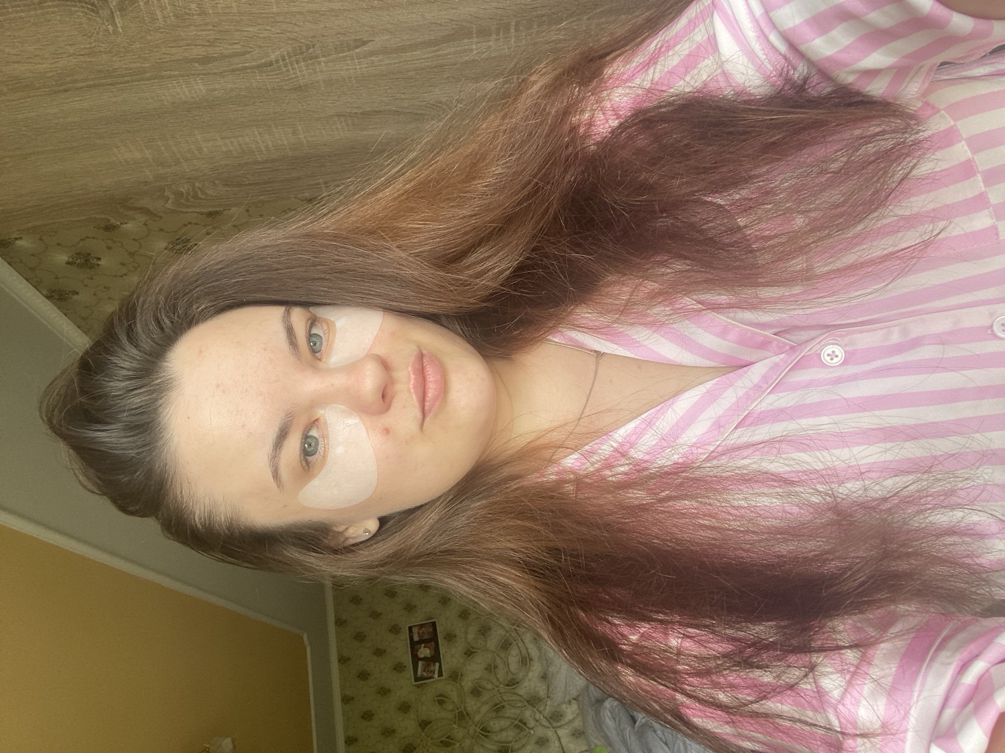 A young woman with long, brown hair and blue eyes is shown in a close-up selfie. She is wearing a pink and white striped pajama top and has white under-eye patches on her face. Her hair is partially obscuring her face and shoulders. The background appears to be an indoor setting, possibly a bedroom, with wallpaper and wooden paneling visible. The lighting suggests it is daytime, with natural light coming from a window or overhead fixture. There is no specific indication of her location in Poltava, Ukraine, beyond the user's prompt.