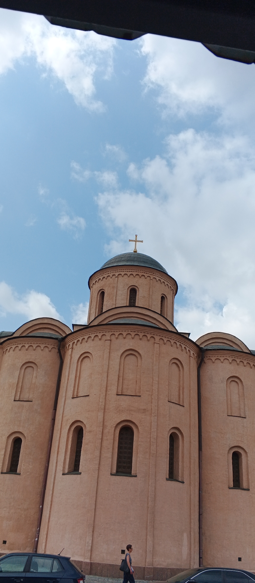 The image captures a portion of the Saint Sophia Cathedral in Kyiv, Ukraine, under a partly cloudy sky. The cathedral's characteristic pale orange facade and dark green dome are visible. A golden cross adorns the top of the dome. The building features multiple rounded arches, some of which appear to be windows. In the foreground, a dark-colored car is parked to the left. To the right of the car, a woman with short dark hair and sunglasses is walking from left to right. She is wearing a dark top and dark pants and carrying a dark bag over her shoulder. The ground beneath the cathedral appears to be paved.