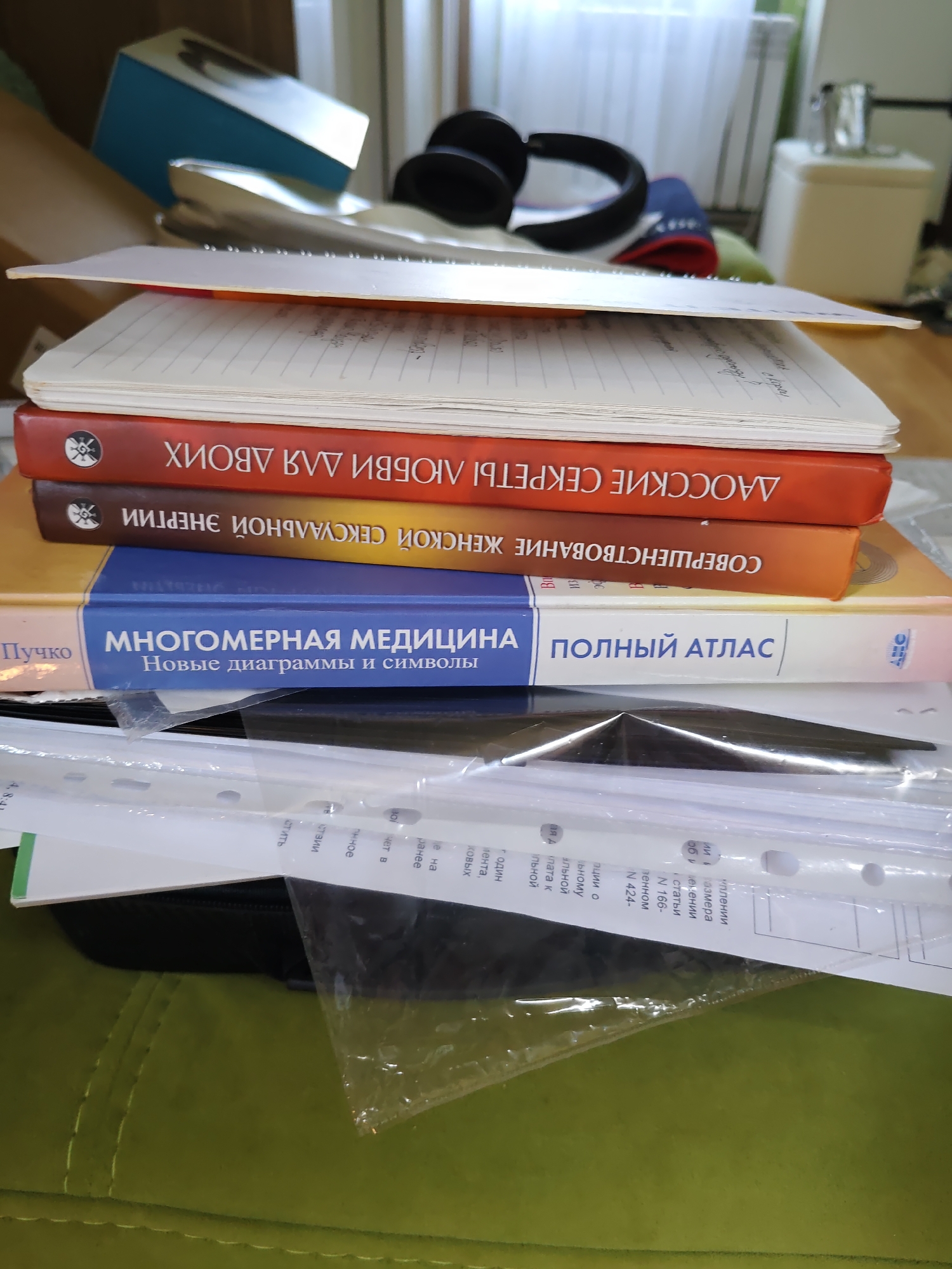 This is a close-up, eye-level, indoor photograph of a stack of books and papers on a green, tufted surface, likely a couch or chair. The scene is cluttered and appears to be in a home or office environment.
The books are stacked horizontally, with the spine of the top book facing away from the viewer. The visible book titles are in Russian and include "ЛАОССКИЕ СЕКРЕТЫ" (LAO SECRETS), "СОВЕРШЕНСТВОВАНИЕ" (IMPROVEMENT), and "МНОГОМЕРНАЯ МЕДИЦИНА" (MULTIDIMENSIONAL MEDICINE) with the subtitle "Новые диаграммы и символы" (New diagrams and symbols). Another book visible has the title "ПОЛНЫЙ АТЛАС" (COMPLETE ATLAS). The book covers are primarily red, orange, yellow, and blue.
Above the books, several sheets of paper are visible, some appearing to be notebook pages with handwritten text or print. To the left and slightly behind the books, a light blue box and a pair of black headphones are partially visible.
Below the books, a black binder or folder is present, and on top of that, clear plastic sleeves containing papers with printed text in Russian are spread out. The text on these papers is small and appears to be from a list or document.
The background is out of focus but shows a window with vertical blinds and a radiator, suggesting an indoor setting. The lighting is soft and natural, likely from the window.
The image does not contain any discernible elements that would allow for specific identification of its location within Rostov-on-Don, Russia. The items and environment depicted are generic and could be found in many indoor settings.