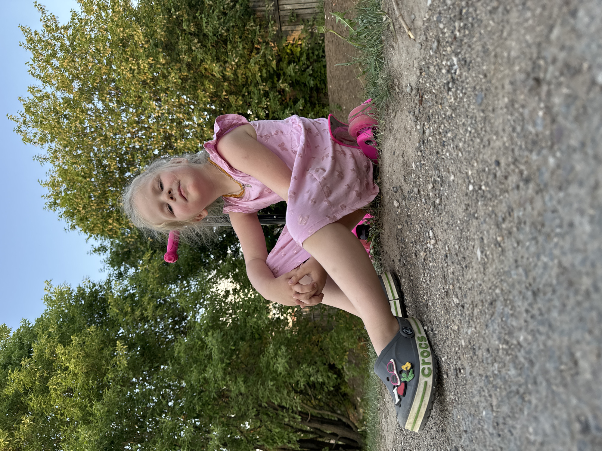 A young girl with blonde hair, wearing a pink dress and grey Crocs with decorative pins, is sitting on a gravel surface. She is looking at the camera. Behind her is greenery, likely trees, and a fence in the background. The photo appears to be taken outdoors in a location with natural light. It is not possible to confirm the location is Горный, Russia from the given information.