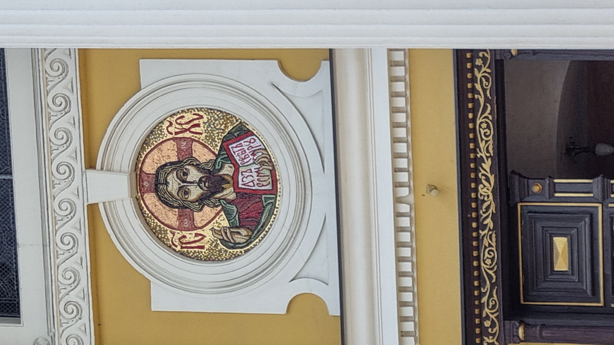 The image shows an outdoor architectural detail on a building in Uzhhorod, Ukraine. Dominating the frame is a circular mosaic depicting Jesus Christ. The mosaic is set within a white, ornate frame against a yellow wall. Jesus is shown with a beard, wearing a green robe over a red garment. He is holding an open book in his left hand, and his right hand is raised in a blessing gesture. Cyrillic inscriptions are visible on the book and within the halo behind Jesus' head. The mosaic is framed by decorative white architectural elements, including scrollwork along the top. Below the mosaic, a dark wooden doorway with gold trim and a patterned door is visible. A white fluted column is to the right of the mosaic. The overall impression is one of religious iconography on a building's facade.