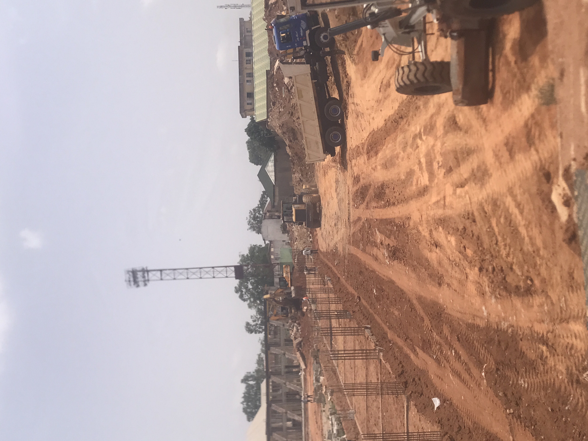 The image shows a construction site in Jos, Nigeria. A partially constructed building with steel rebar framework is visible in the foreground. An unpaved, reddish-brown dirt road cuts through the center of the scene, with heavy machinery including a dump truck, loader, and excavator present. A communications tower stands in the background against a hazy sky. The environment appears dusty, with buildings and trees visible in the midground.