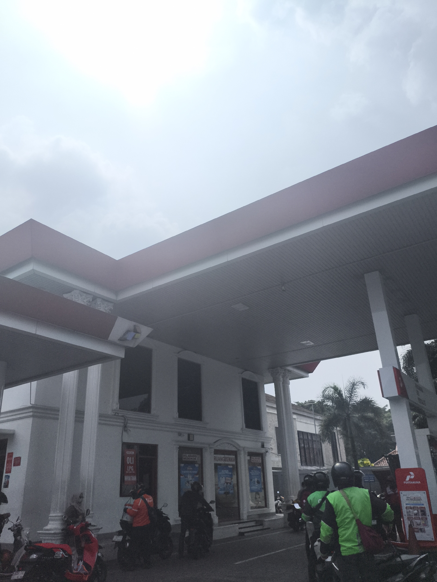 The image depicts a street scene at a gas station in Depok, Indonesia. The sky is overcast. The foreground features several motorcycles, with riders wearing helmets. Some riders are wearing bright green jackets, indicating they may be delivery personnel. One motorcycle is red, while others are darker in color. The gas station has a white building with a red-trimmed roof and a covered canopy. Signs are visible on the building, including one that says "OLI LPG". In the background, palm trees and other buildings are visible. The overall impression is a busy urban environment.