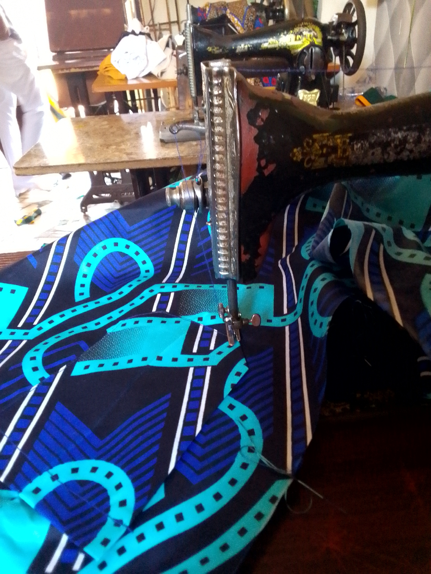 The image shows a close-up of a vintage sewing machine stitching a vibrant patterned fabric. The fabric is predominantly dark, featuring a design of turquoise and blue shapes, including square-dotted lines and geometric patterns with white stripes. The sewing machine, showing signs of age with worn paint, has a metallic needle bar actively engaged with the fabric. In the background, there are glimpses of a table and other objects, suggesting an indoor setting, possibly a workshop or home environment. The location is Unknown, Unknown.