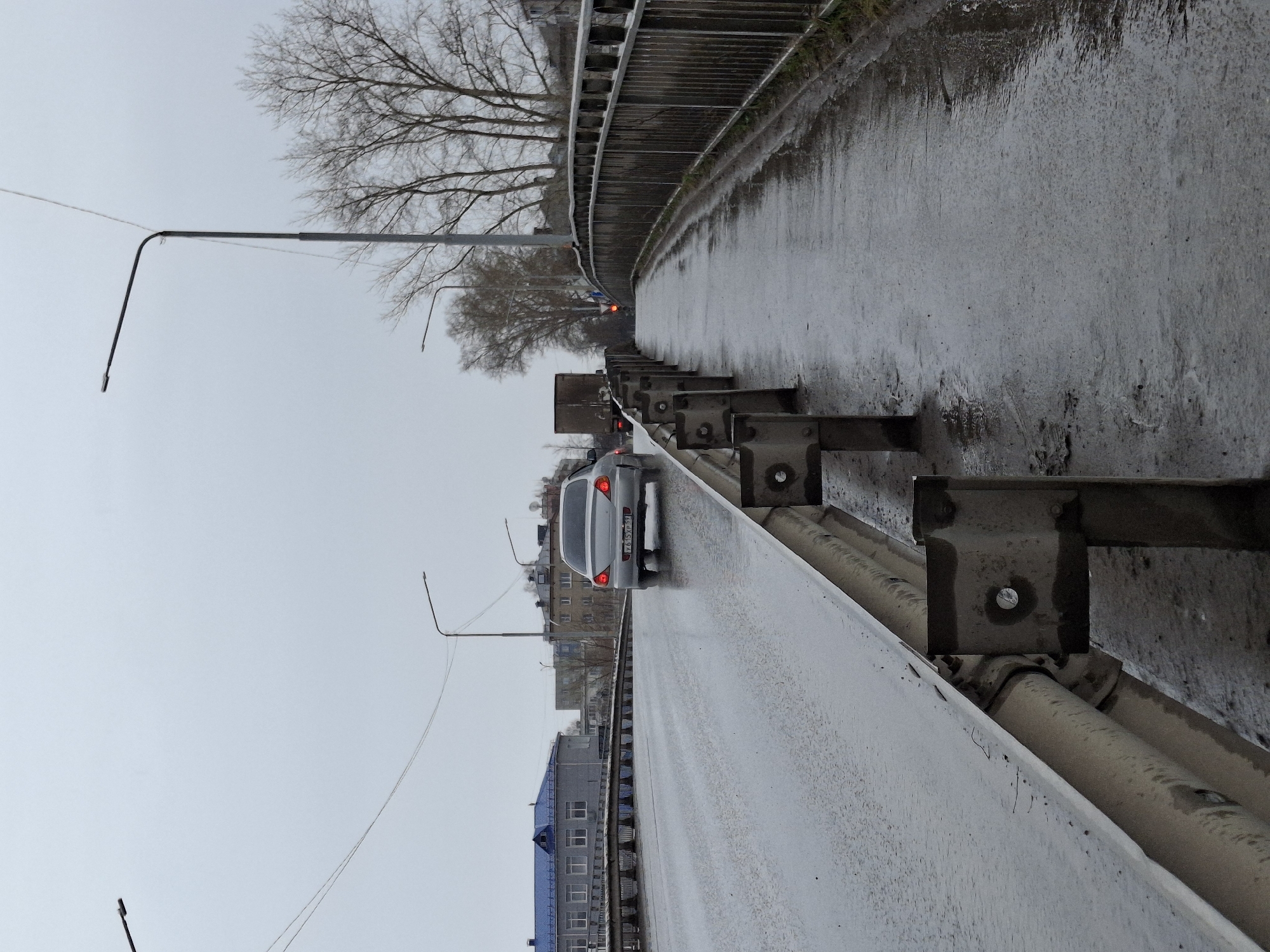 The image depicts a wet, overcast day on a bridge or overpass in what appears to be the city of Chaikovsky, Russia, based on the visible license plate. A light-colored sedan is driving on the road, with its brake lights illuminated. The road is flanked by guardrails on both sides. The guardrail closer to the camera is a combination of a concrete barrier and metal rails with visible mounting hardware. Beyond the guardrail on the far side of the road, buildings and trees are visible. Streetlights line the road, and a single red light is also visible in the distance. The overall scene suggests a gray and damp urban environment.