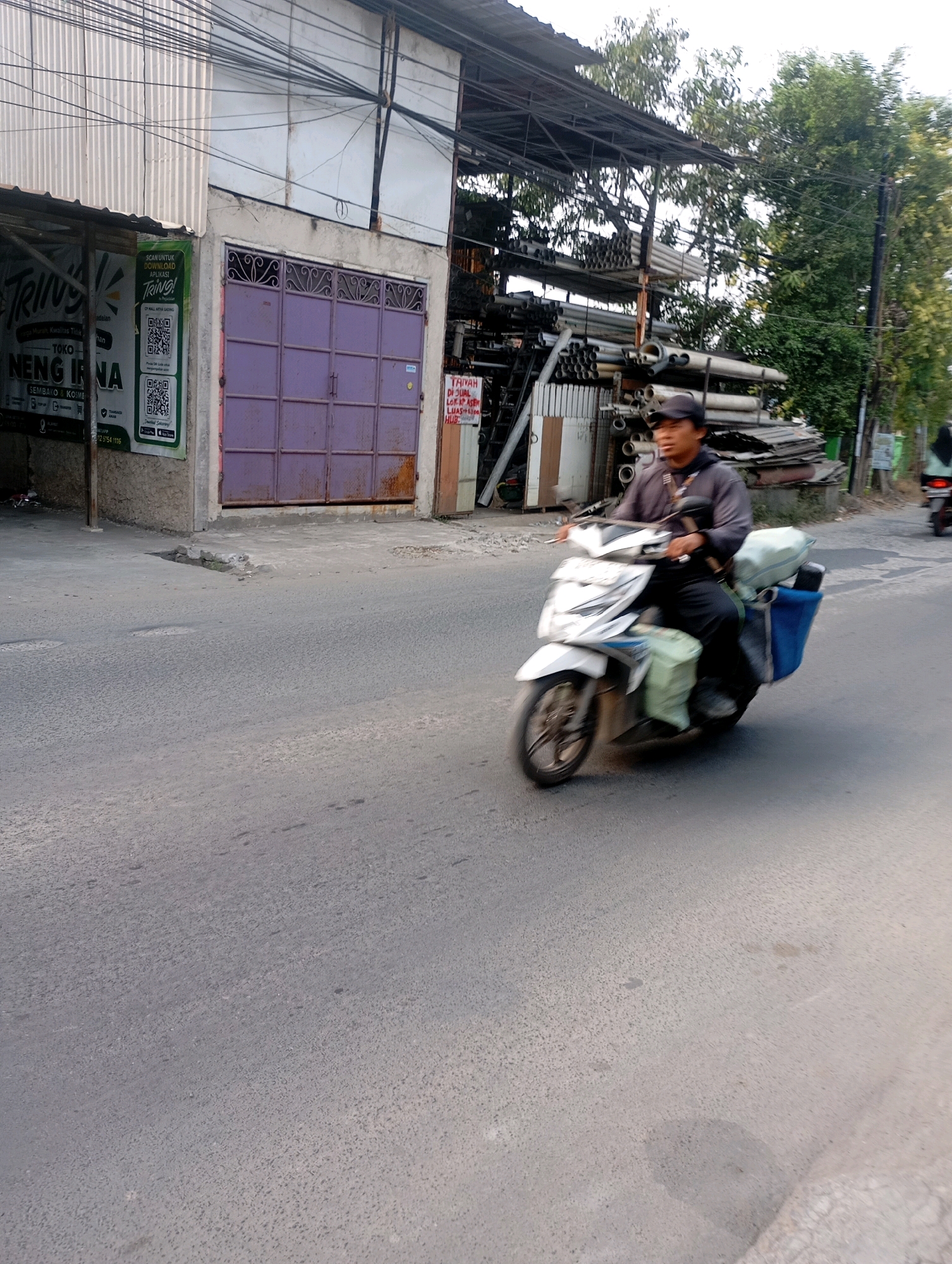 A man wearing a black cap and a dark gray long-sleeved shirt is riding a white scooter on an asphalt road in Babelan, Indonesia. The scooter has a blue basket on the back and green bags hanging on the sides. The man is looking to the left and appears to be in motion. In the background, there is a building with a purple garage door and corrugated metal siding. Shelves filled with pipes are visible next to the building. Trees and power lines are also present in the scene. The road surface is rough and appears to be a bit dusty.