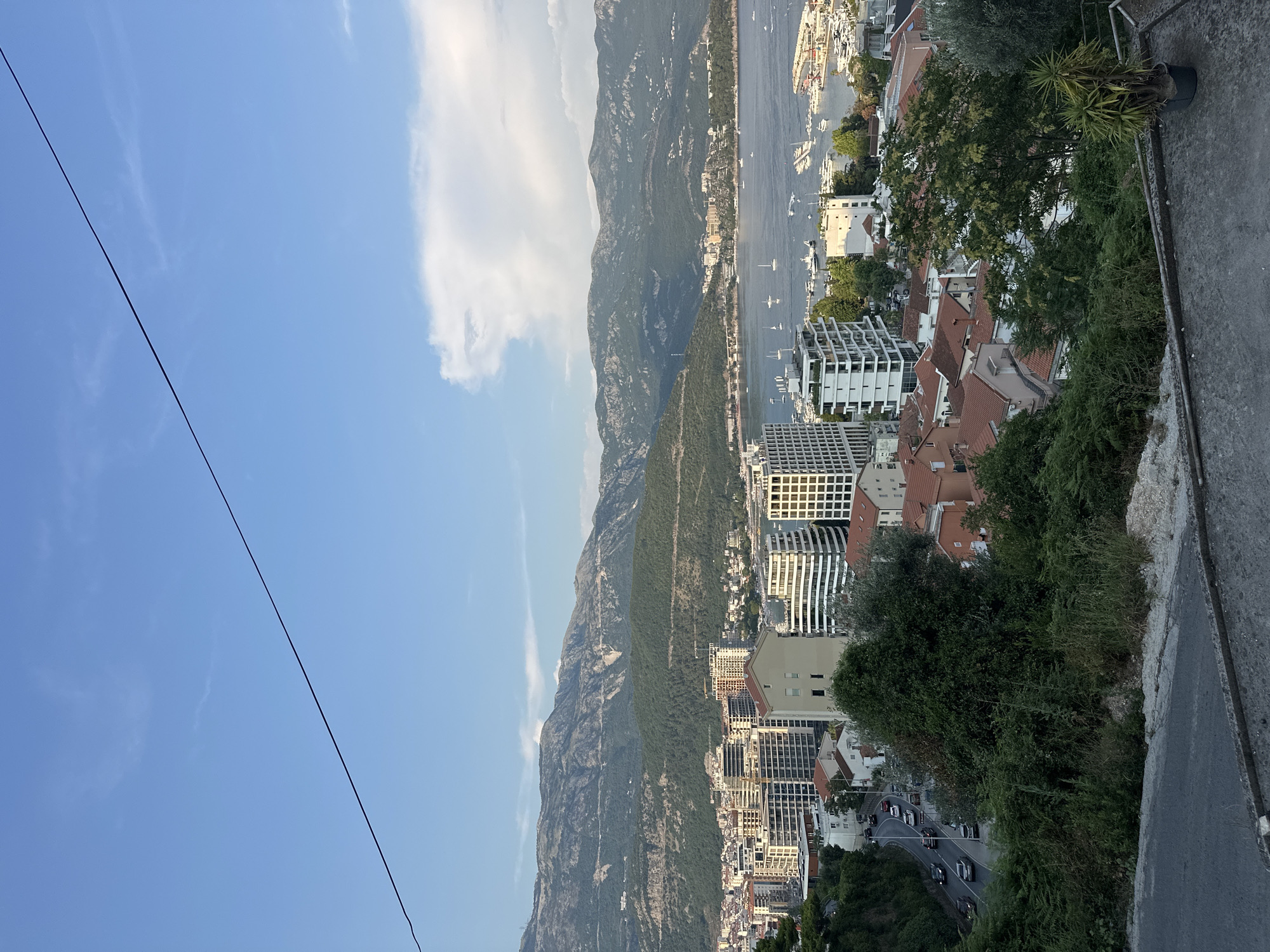 This is a vertical, eye-level shot of a coastal city with a mountain backdrop. The scene is dominated by the dramatic contrast between a rugged, green mountain range and a developed urban area.
The urban area is densely packed with buildings of varying architectural styles. Modern, white buildings with distinctive curved facades stand alongside older structures with red-tiled roofs. The buildings are clustered along the coastline, with a harbor visible in the distance, dotted with numerous sailboats and yachts.
The mountain range is a prominent feature, covered in dense green vegetation, with rocky outcrops visible in places. The sky is a clear, bright blue, with a few wispy white clouds. The sun appears to be shining, casting a warm light over the scene, suggesting it is daytime, likely late afternoon due to the soft light.
A winding road with several cars can be seen on the right side of the image, descending towards the city. Lush green trees and foliage are abundant, growing on the slopes and within the urban landscape. A thin black wire cuts diagonally across the sky from the lower left to the upper right of the frame.
The overall impression is one of a beautiful, vibrant coastal city nestled between impressive natural scenery. The location is identified as Budva, Montenegro. There is no visible text in the image.