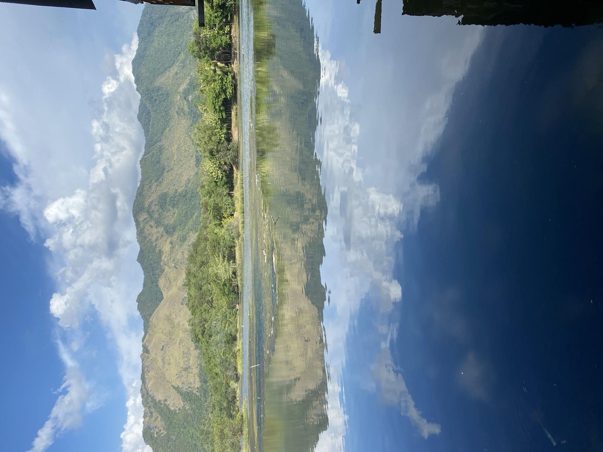 The image depicts a serene landscape with a body of water in the foreground, reflecting a blue sky dotted with white clouds and a distant mountain range. The mountains are covered in green vegetation, with some areas appearing lighter, possibly due to dry grass. Trees and foliage line the shore of the water. Part of a wooden structure is visible on the right side of the image, casting a shadow on the water. The water itself is calm, with some visible debris floating on its surface. The overall impression is one of natural tranquility. While the image is described as being in Aneuk Paya, Indonesia, specific geographical landmarks within the city are not identifiable from the visual information provided.