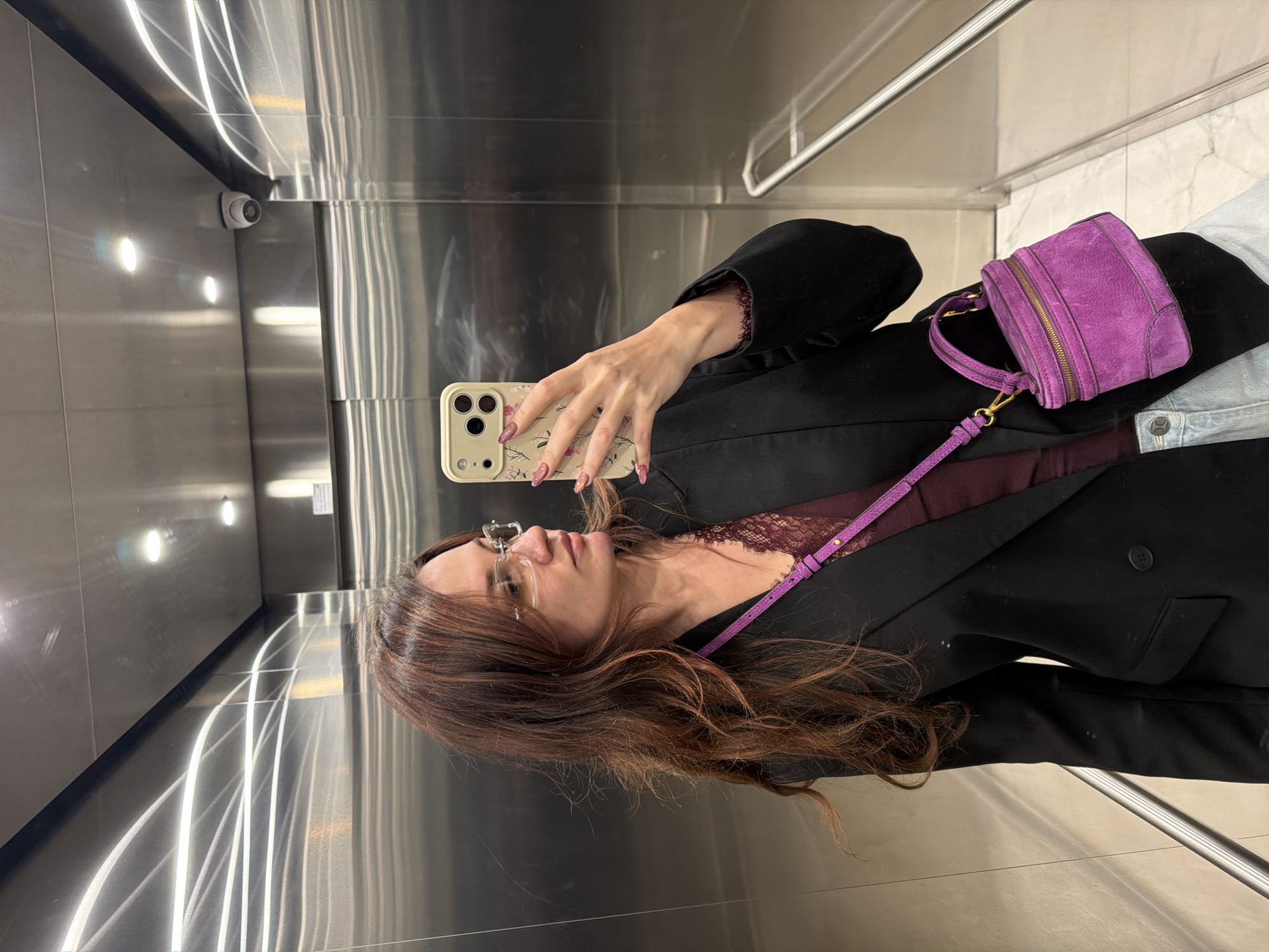An individual is captured taking a mirror selfie in a highly reflective, metallic environment, likely an elevator or a modern hallway, located in Belgrade, Serbia. The person, viewed from the chest up, is holding a light-colored smartphone with a floral-patterned case in their right hand, angled to photograph their reflection. Their nails are manicured with a pinkish-purple polish and design.
The person has long, wavy, reddish-brown hair and is wearing clear-framed glasses. They are dressed in a black blazer over a deep purple or maroon lace top, with light blue jeans visible at the bottom right. A vibrant purple suede crossbody bag with gold hardware hangs from their left shoulder.
The setting features brushed metal walls and ceiling, which create numerous bright, elongated reflections of recessed overhead lights. A small, white security camera is visible in the upper left corner of the reflection. A silver handrail is also reflected horizontally across the lower part of the scene. The lighting is artificial and bright, suggesting an indoor setting. There is no discernible text visible in the image.