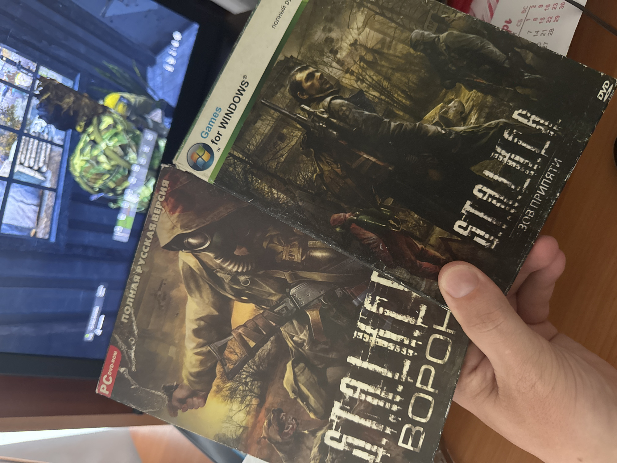 A person is holding two physical PC game cases, "S.T.A.L.K.E.R.: Call of Pripyat" and "S.T.A.L.K.E.R.: Clear Sky" ("S.T.A.L.K.E.R.: Вороны" - literal translation "Crows", a Russian version of Clear Sky). The "Call of Pripyat" case has a "Games for Windows" logo. The background shows a computer monitor displaying a video game. The setting appears to be indoors, on a desk, perhaps. Also, there's a calendar on the desk. Location: Chuprino, Russia not deducible from the image.
