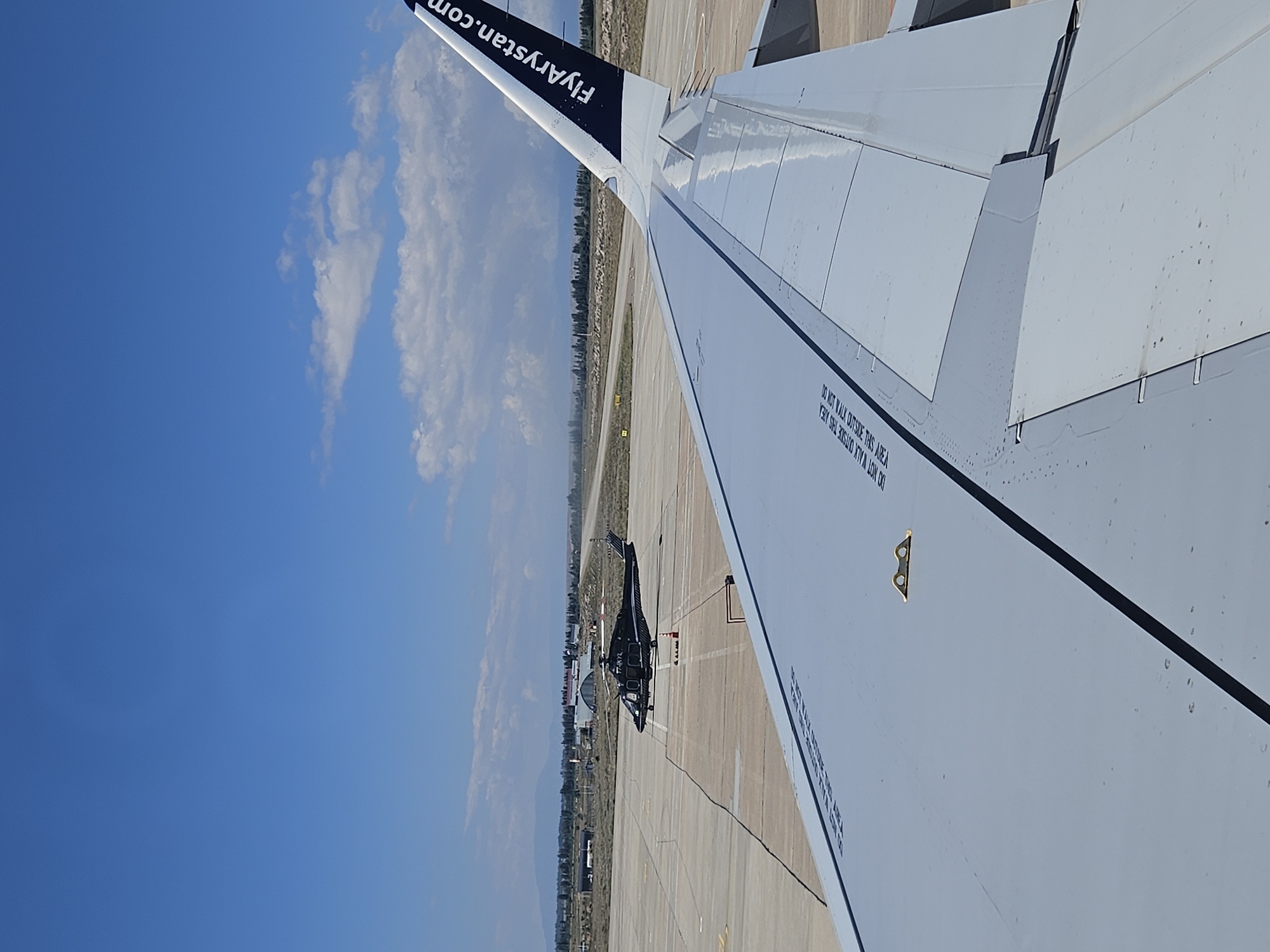 The image captures an airfield scene under a clear blue sky with scattered clouds. In the foreground, the wing of a passenger airplane occupies the lower right portion of the frame, displaying the text "FlyArystan.com" on its tail fin, which extends upwards. On the tarmac, a black helicopter with the registration "T7-HKYL" is parked. The background reveals airport buildings, a runway, and a treeline, with distant mountains under the horizon. The environment appears to be sunny and dry, consistent with an airport in Choktal, Kyrgyzstan. There are no people visible in the image.